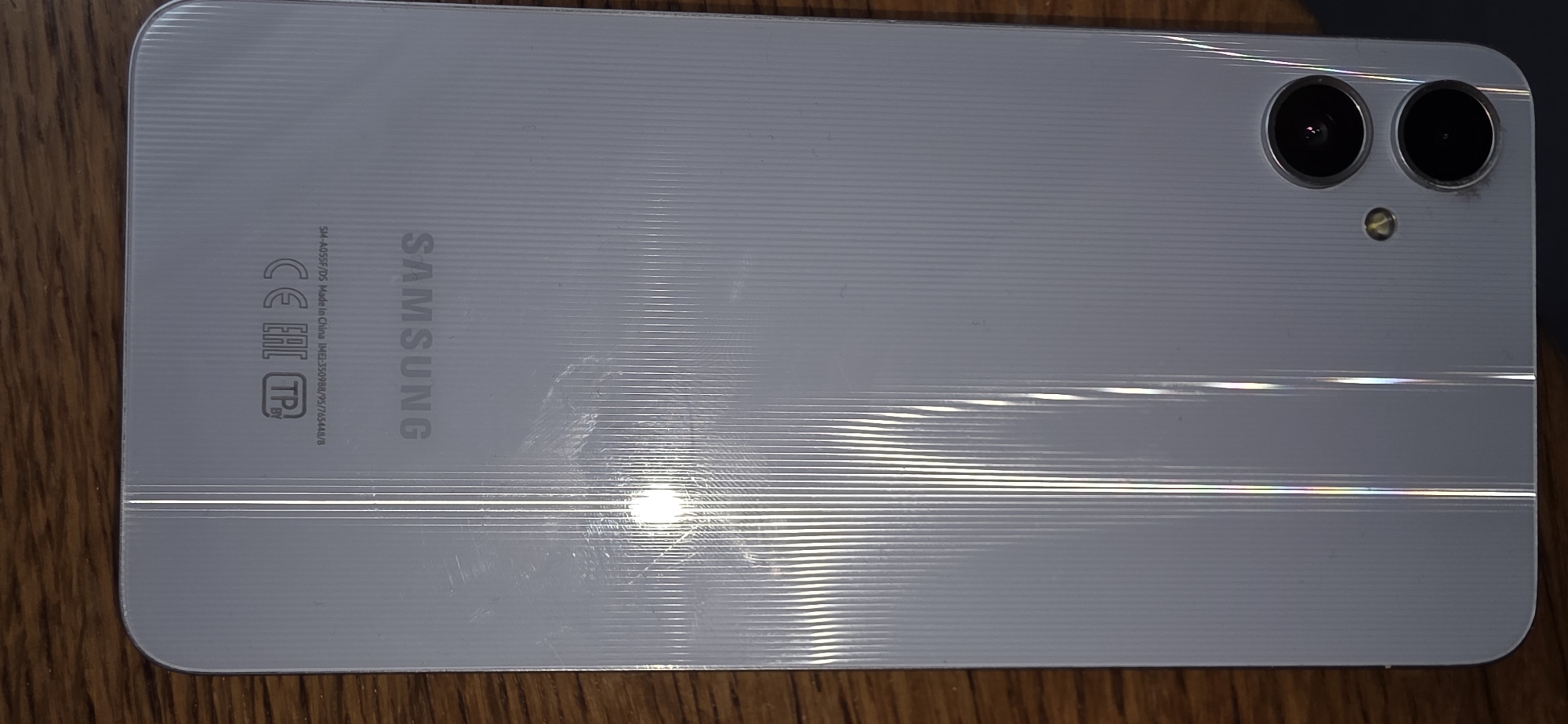 The image shows the back of a white Samsung smartphone resting on a wooden surface. The phone has a textured finish with vertical lines and features a dual-camera system on the lower right side. There are some scuff marks and smudges on the phone's surface, and a bright light reflection is visible in the upper left quadrant. Textual markings on the phone include "SAMSUNG" logo, "CE EAC TP BY", and "SM-A055F/DS Made in China IMEI:350988/75/65448/8". The background is a wooden surface with visible grain. There are no people or any other objects or activities depicted in the image. There is no indication of the location being in Marum, Netherlands.