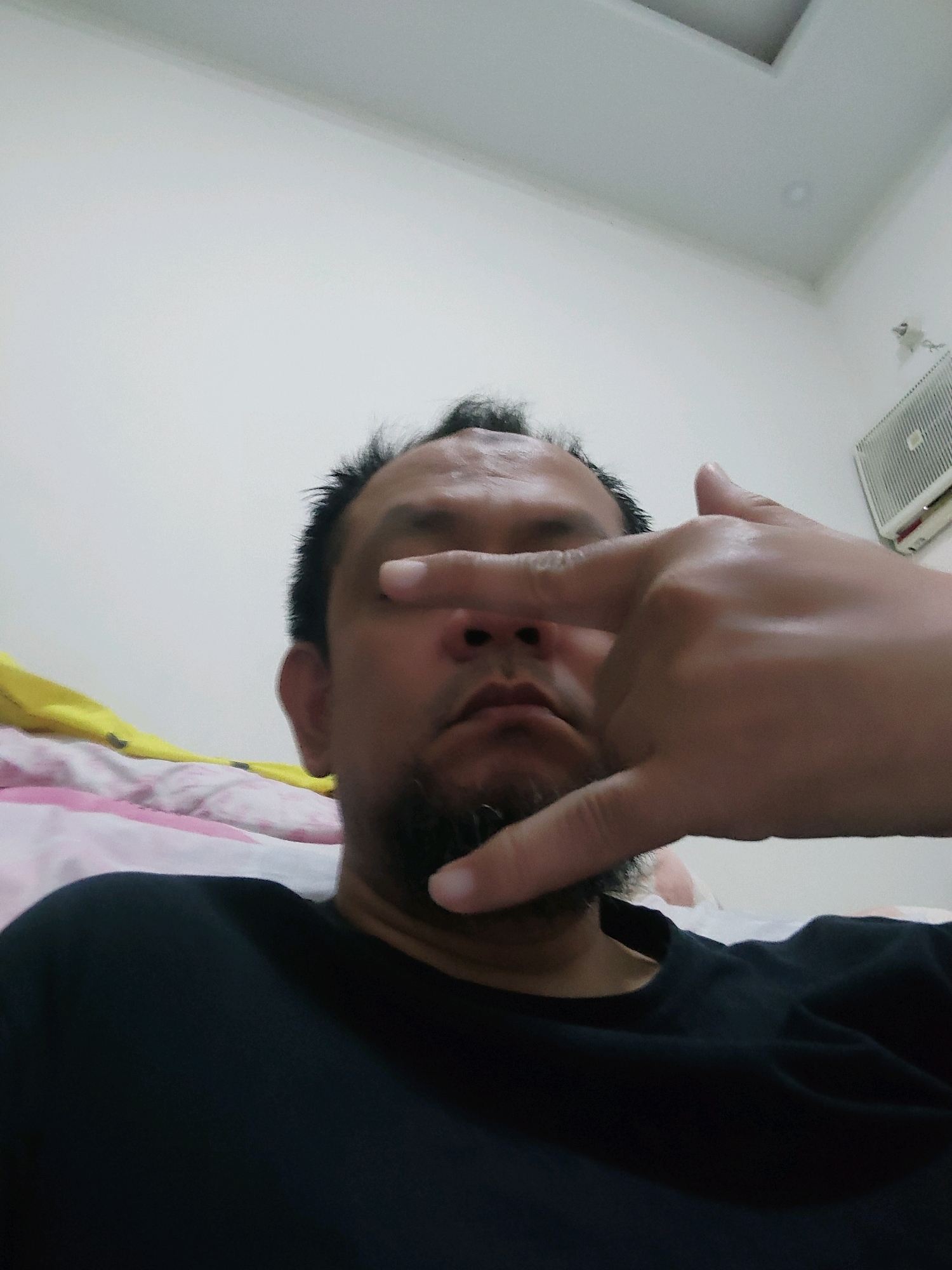 The image is a close-up, low-angle selfie taken indoors. A person's face is partially obscured by their hand, which is held up to their eyes. The person has dark hair, a goatee, and is wearing a black t-shirt. They appear to be lying down on a bed with pink and white striped bedding. In the background, a white wall is visible, along with a cream-colored ceiling and a white ventilation unit mounted on the wall. The overall environment suggests a domestic setting. There is no visible indication of the location being in Bogor, Indonesia.