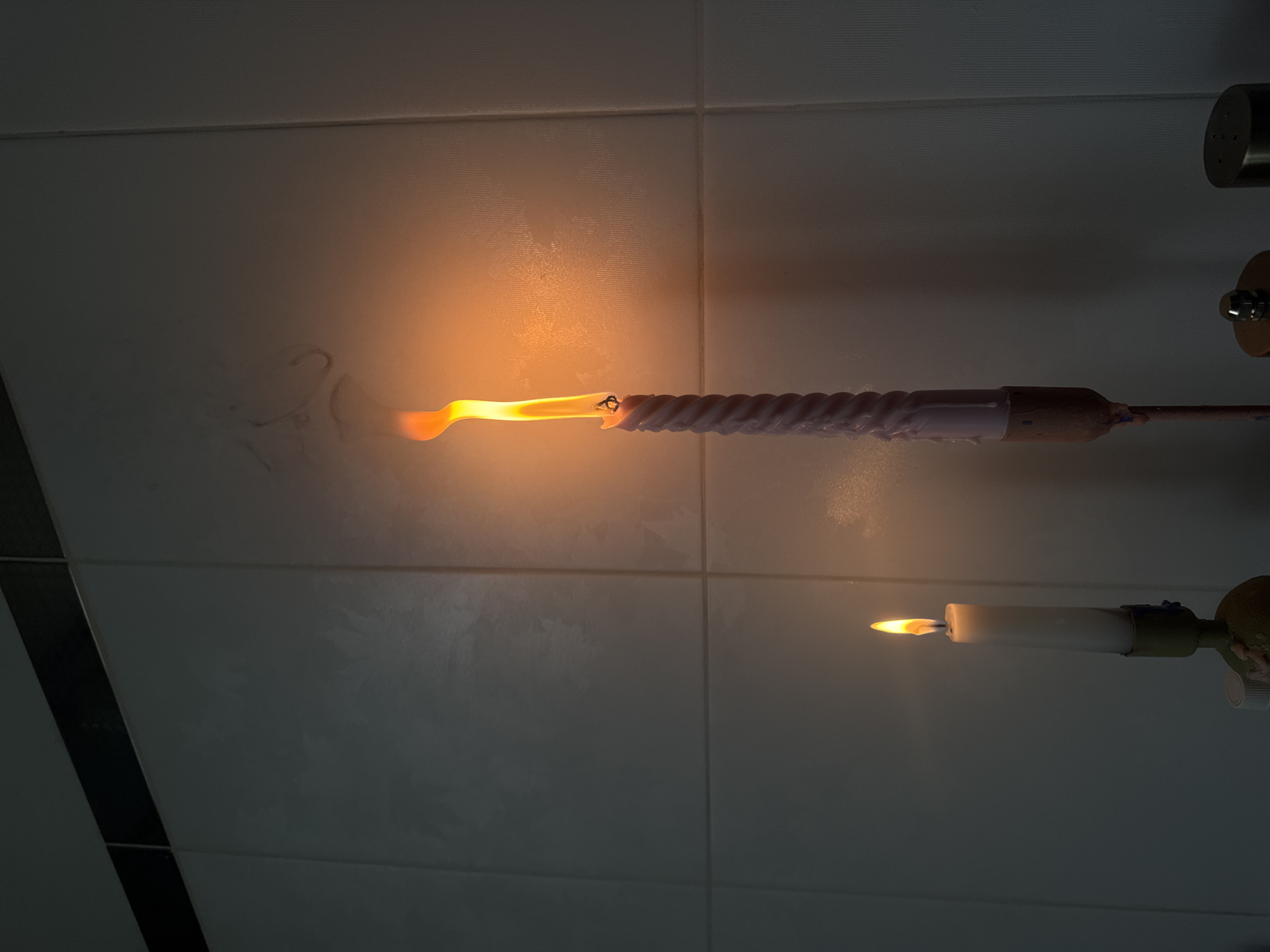 The image displays two burning candles horizontally mounted against a tiled wall in a dimly lit indoor setting, likely in Moscow, Russia. The upper candle is a long, twisted, light purple or off-white wax candle, held by a thin rod extending from the right. Its flame is large, elongated, and bright orange-yellow, illuminating the immediate wall area. Swirls of dark smoke are visible on the light-colored tiles above and to the left of this flame.
Below it, a shorter, white candle also burns horizontally, secured by a metallic fixture that appears to be part of a wall-mounted apparatus, possibly with green or brown accents. Its flame is smaller and more contained. The background consists of light-colored, possibly white or light gray, square tiles with dark grout lines forming a grid pattern. Darker, possibly black, vertical tile strips are visible on the far left. On the far right, near the top, a metallic cylindrical object with small holes is partially visible, along with another metallic fixture below it. The low light conditions, with candles as the primary light source, suggest an evening or nighttime scene. No people or text are visible.