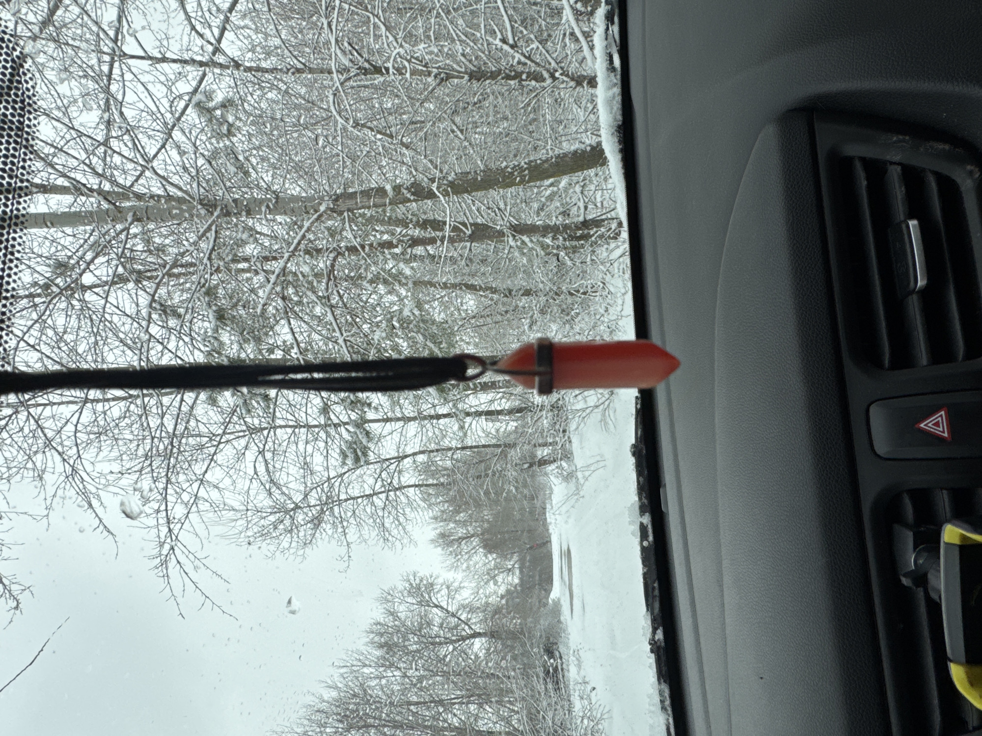 The image shows a view from inside a vehicle, likely a car, looking out onto a snow-covered scene. Bare trees, heavy with snow, fill the background. In the foreground, hanging from the rearview mirror or dashboard, is an orange, pointed crystal pendant on a black cord. To the right of the frame is part of the vehicle's interior, including an air vent and the hazard light button. The specific location in Chaikovsky, Russia, cannot be determined from the image alone.