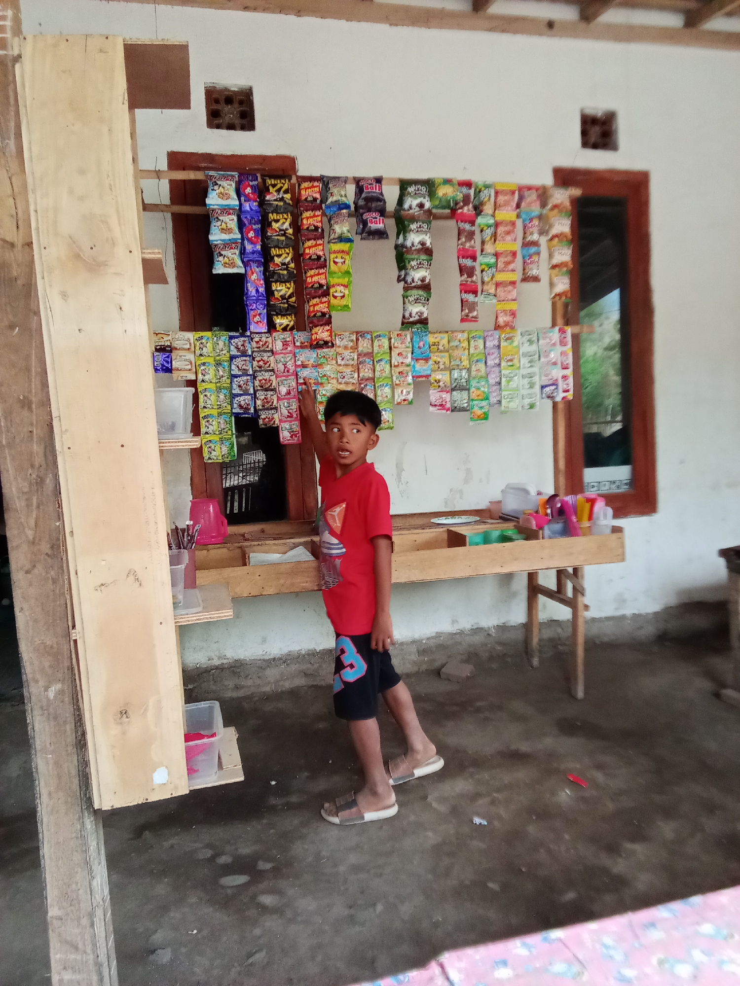A young boy stands in front of a small, makeshift store in Trebungan, Indonesia. The store is stocked with various small, colorful snack packets, hung on wooden racks attached to a wall. Some packets are arranged in rows on a wooden table beneath the racks. The boy, wearing a red t-shirt and dark shorts, is pointing towards the snack packets. To the left of the store, a tall wooden structure, possibly a shelf or a wall, stands precariously. The floor is made of concrete, and the background shows a white wall with a window and a small ventilation brick. The overall scene depicts a humble, rural retail setup.
