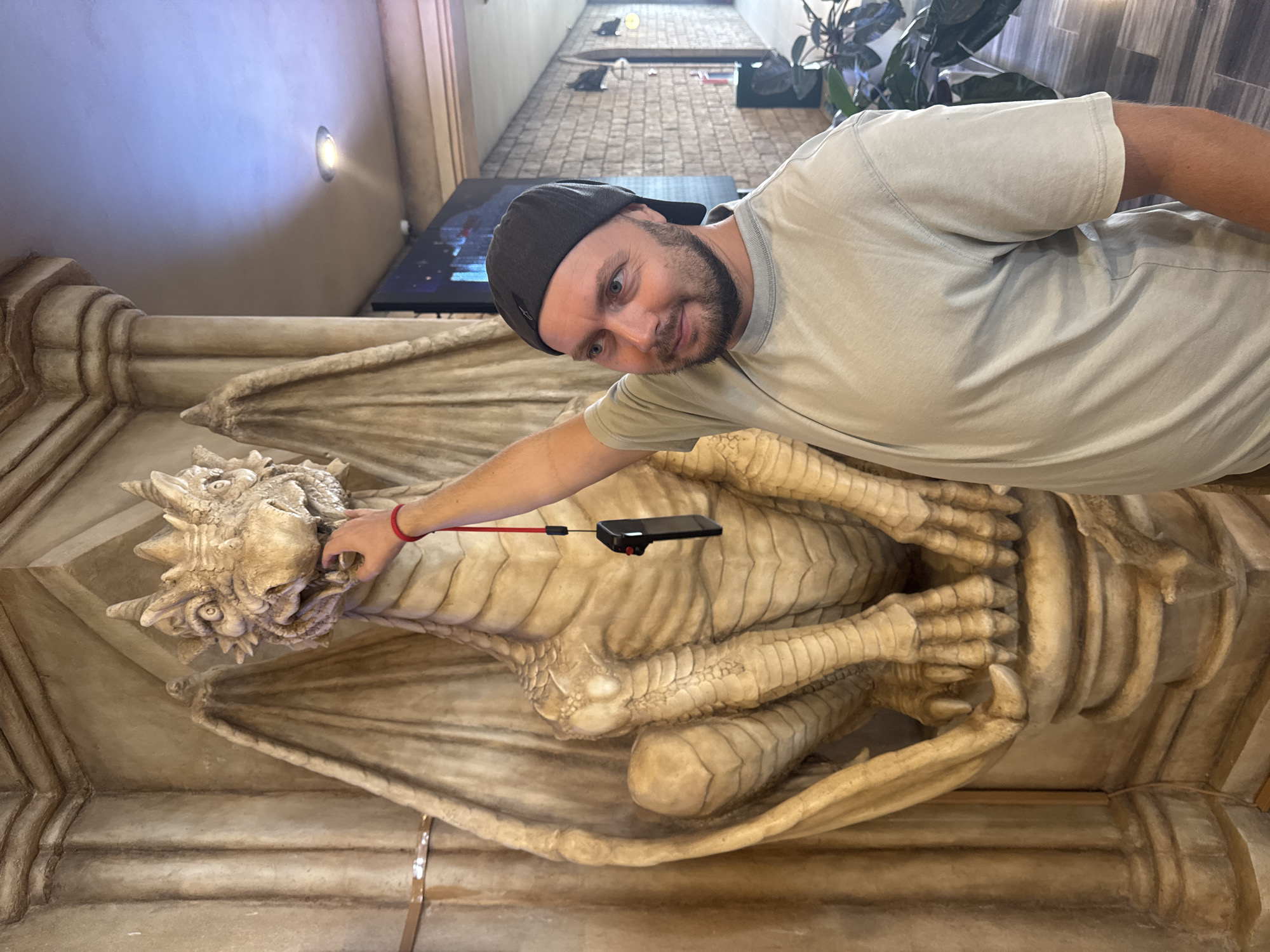 A man wearing a gray t-shirt and a dark baseball cap is posing with a dragon statue in an indoor setting. The statue, detailed with wings and scales, appears to be made of a light-colored material, possibly plaster or resin. The man is touching the dragon's head with his right hand, while holding a small electronic device with a red cord attached near the dragon's chest. The background includes a wall with a recessed light fixture, a television, and a glimpse of a tiled hallway with plants. The location is uncertain but is alleged to be Đà Nẵng, Vietnam.