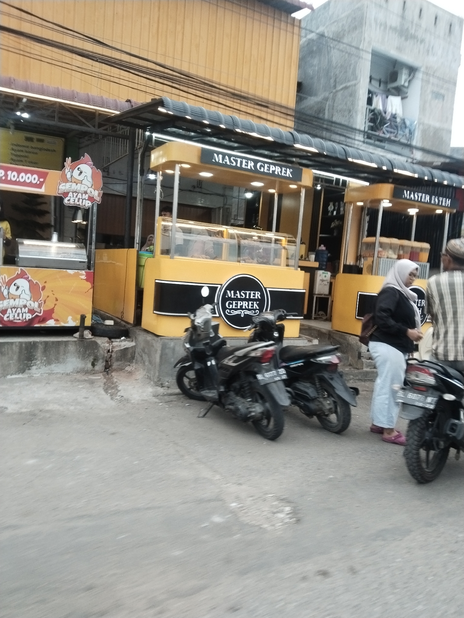 The image shows a street scene in Lhokseumawe, Indonesia, likely in front of food stalls. The primary focus is on two brightly colored food stalls with yellow and white facades, branded "MASTER GEPReK" and "MASTER ESTEH". Both stalls appear to be open for business, with glass display cases. In the foreground, two motorcycles are parked. On the right, a woman wearing a light blue hijab and a dark shirt, and light blue pants, is walking away from the stalls. Another person, partially visible and wearing a striped shirt, is standing next to her. The street is paved with asphalt, and there are some cracks and debris on the ground. In the background, a concrete building with an open window showing clothes hanging to dry is visible, as well as overhead power lines. A sign with the text "P. 10.000,-" and a cartoon chicken graphic is visible on the left side of the frame, suggesting prices for food items. The overall impression is a busy, informal street-side food vendor area.
