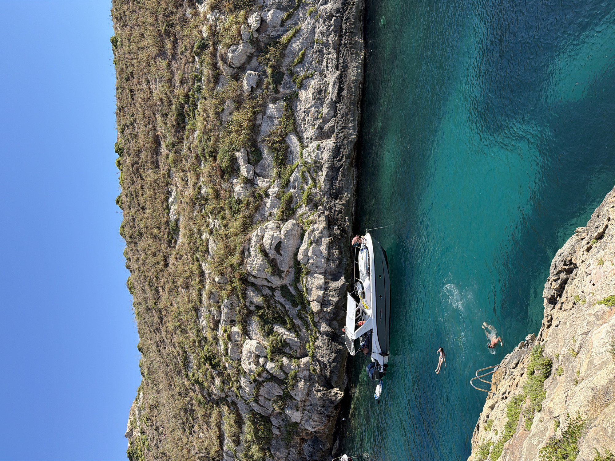 This is a photograph taken in Xlendi, Malta, showing a clear blue sea with a white motorboat anchored in the middle. There are two people swimming in the water, and another person on the boat. The sea is surrounded by rocky cliffs covered with sparse vegetation. The sky is clear and blue.