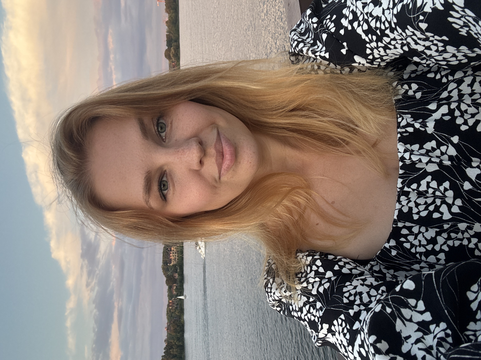 A young woman with blonde hair and freckles, wearing a black and white floral top, takes a selfie on a waterfront with a river and a cloudy sky. The background shows a city skyline with trees and buildings, and boats on the water. The overall mood is relaxed and casual. The location is the city of Zhukovo, Russia, likely during a warm season.