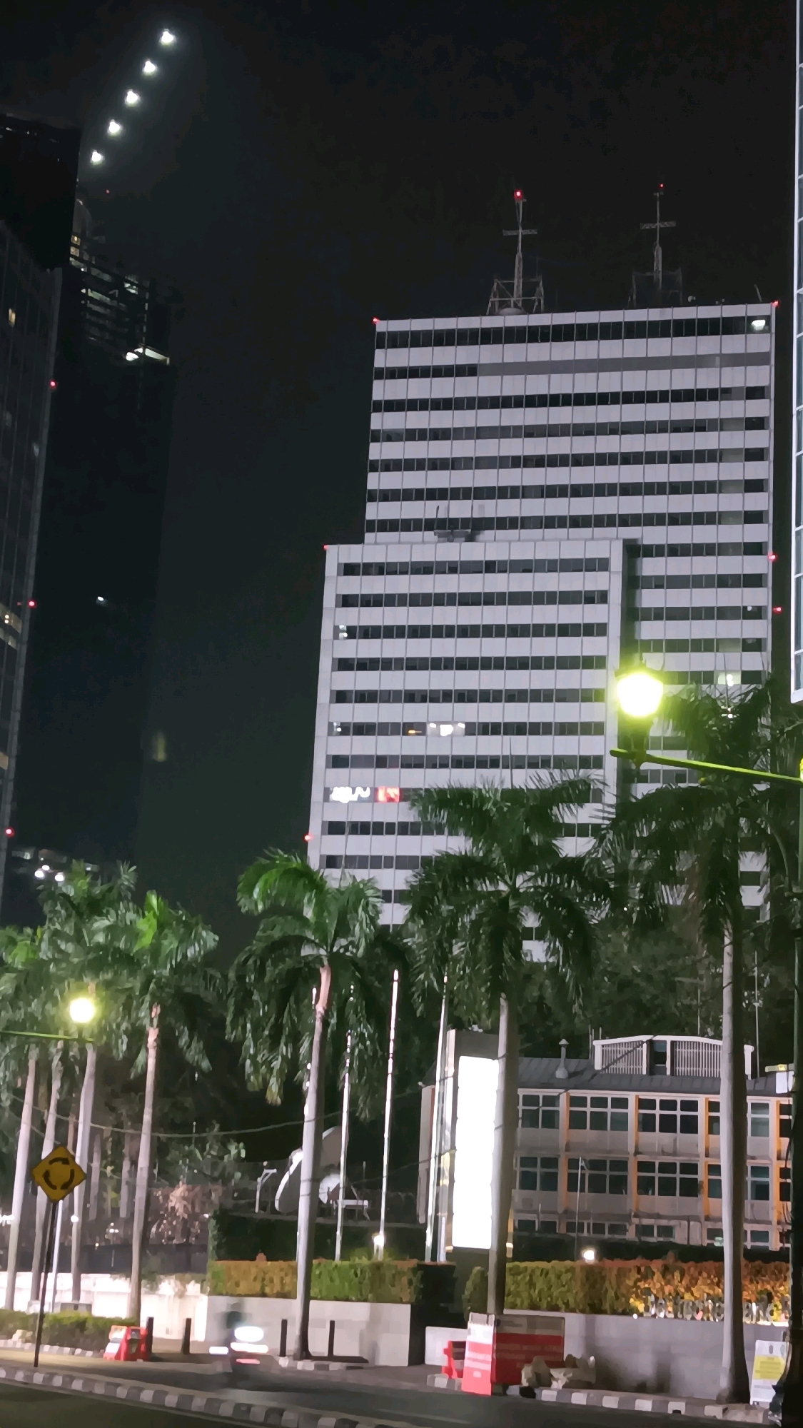 The image depicts a nighttime scene in Central Jakarta, Indonesia, featuring a row of palm trees in the foreground, bordering a street with some traffic cones and barricades. Beyond the trees, a large, white, multi-story building with a grid of windows dominates the center of the frame. Two prominent antennae with red lights are visible on top of this building. To the left of this main building, another, taller, darker skyscraper is partially visible, its upper section illuminated by a series of bright lights against the night sky.
To the right of the main white building, a lower, lighter-colored building with many windows is visible, situated behind some landscaping. A bright streetlamp illuminates the area near the palm trees. The overall atmosphere is urban and modern, typical of a city at night. There is no visible activity of people in the image. The time of day is clearly night, as indicated by the dark sky and the artificial lighting. There are no explicit weather cues.
A partially visible logo or sign on the central white building appears to display the letters "MNN" in red. A yellow circular traffic sign with a black arrow is also visible on a pole to the left of the palm trees.