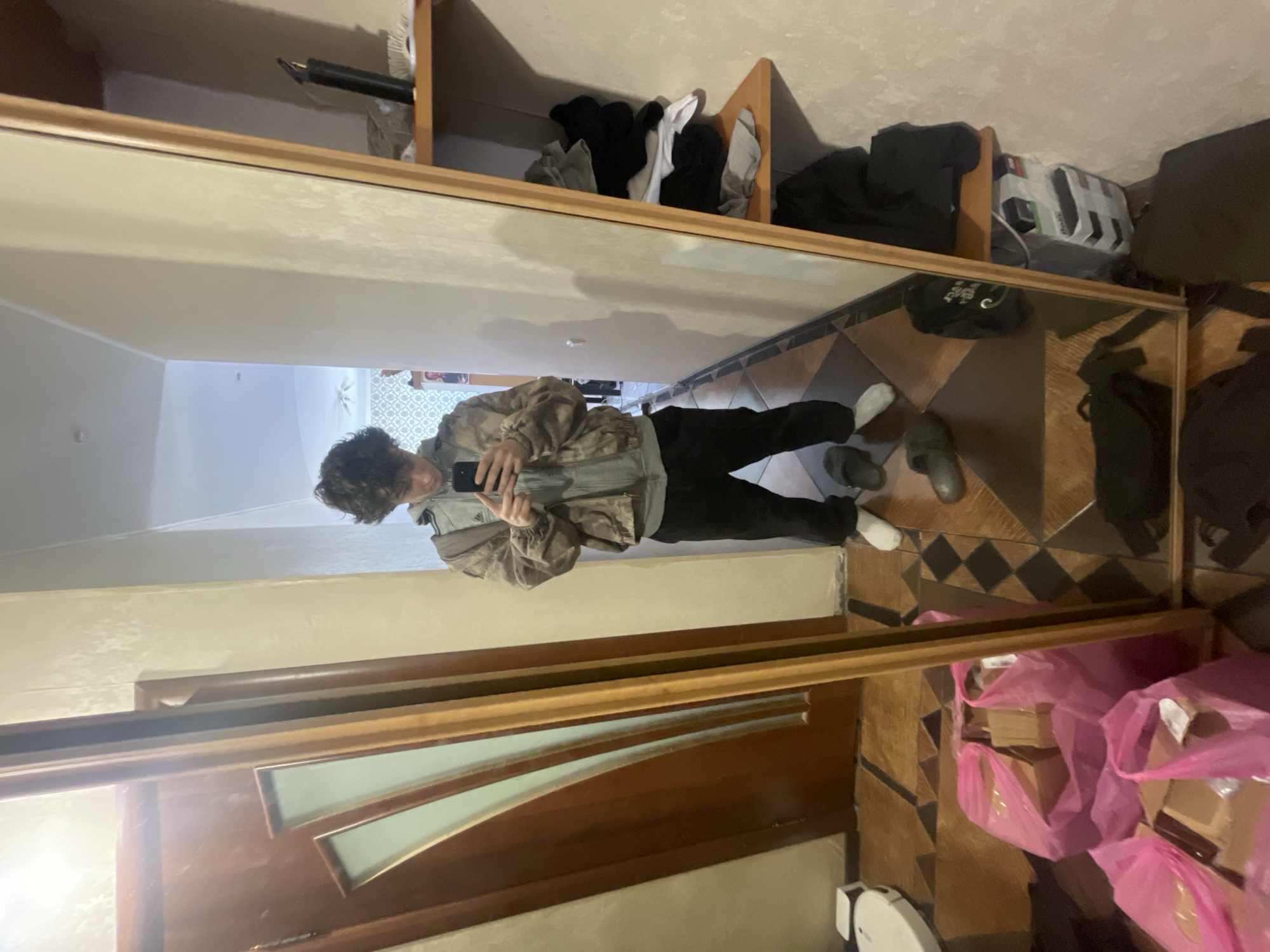 A person is taking a photo of their reflection in a full-length mirror. The person has curly dark hair and is wearing a gray jacket and black pants. They are holding a smartphone in their hands. The mirror reflects a hallway with a decorative tile floor and walls of light beige. Behind the person in the reflection, a glimpse of a brighter room with patterned wallpaper can be seen. In the background of the real room, above the mirror, is a shelf with clothing items and other objects. Below the mirror, there are cardboard boxes in pink plastic bags. A robotic vacuum cleaner is on the floor near the boxes. The location is not discernible.