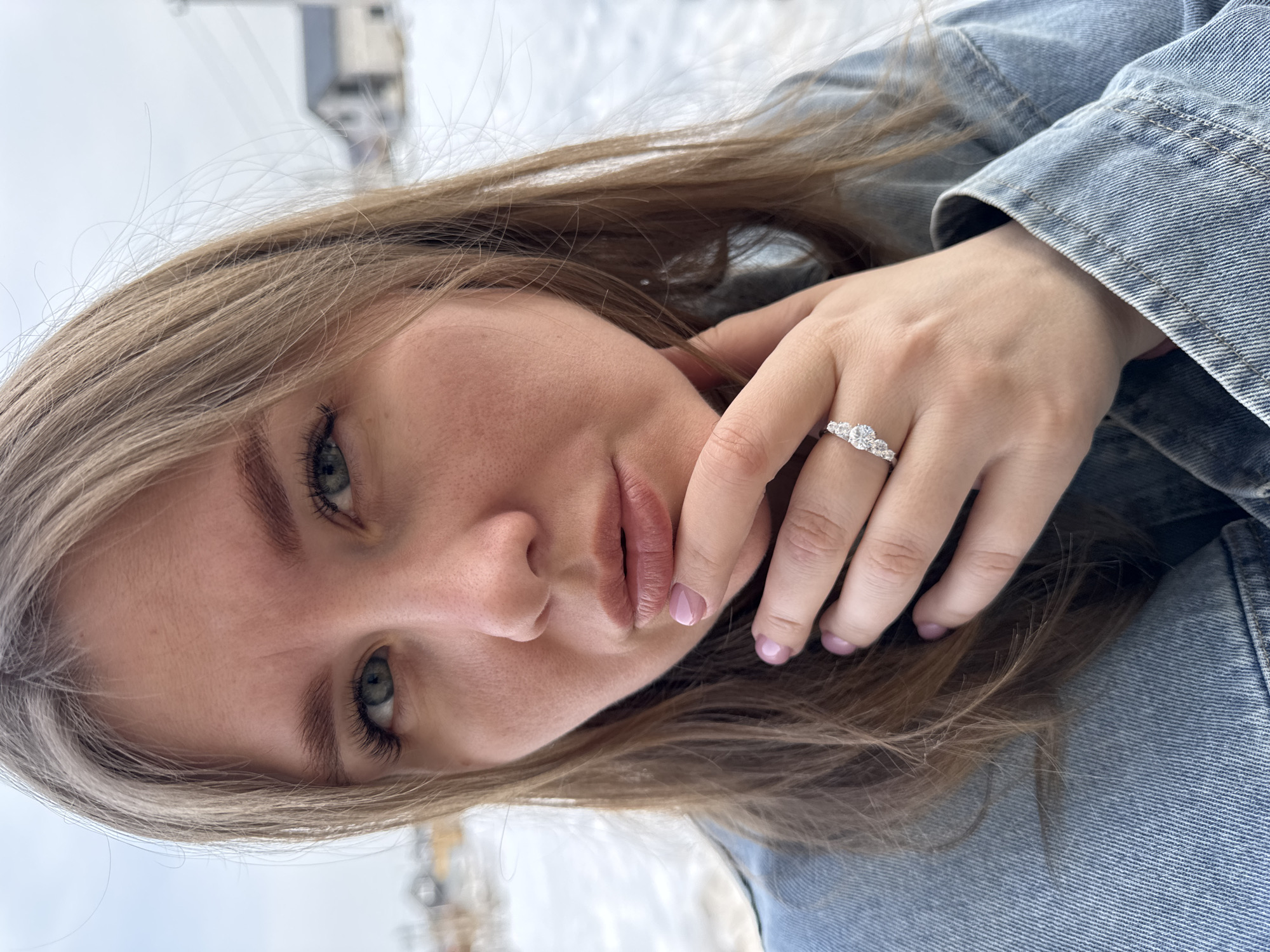 A close-up portrait of a female subject is depicted outdoors in Gornyi (Dvurechenskoe S/P), Russia. The subject has light brown hair, green eyes, and is wearing a light blue denim jacket. Her right hand, featuring light pink nail polish, is positioned with fingertips touching her chin. A silver-colored ring with three clear stones is visible on her right ring finger. The background consists of a blurred winter landscape, showing snow-covered ground, distant residential structures with dark roofs, and utility poles under an overcast sky.