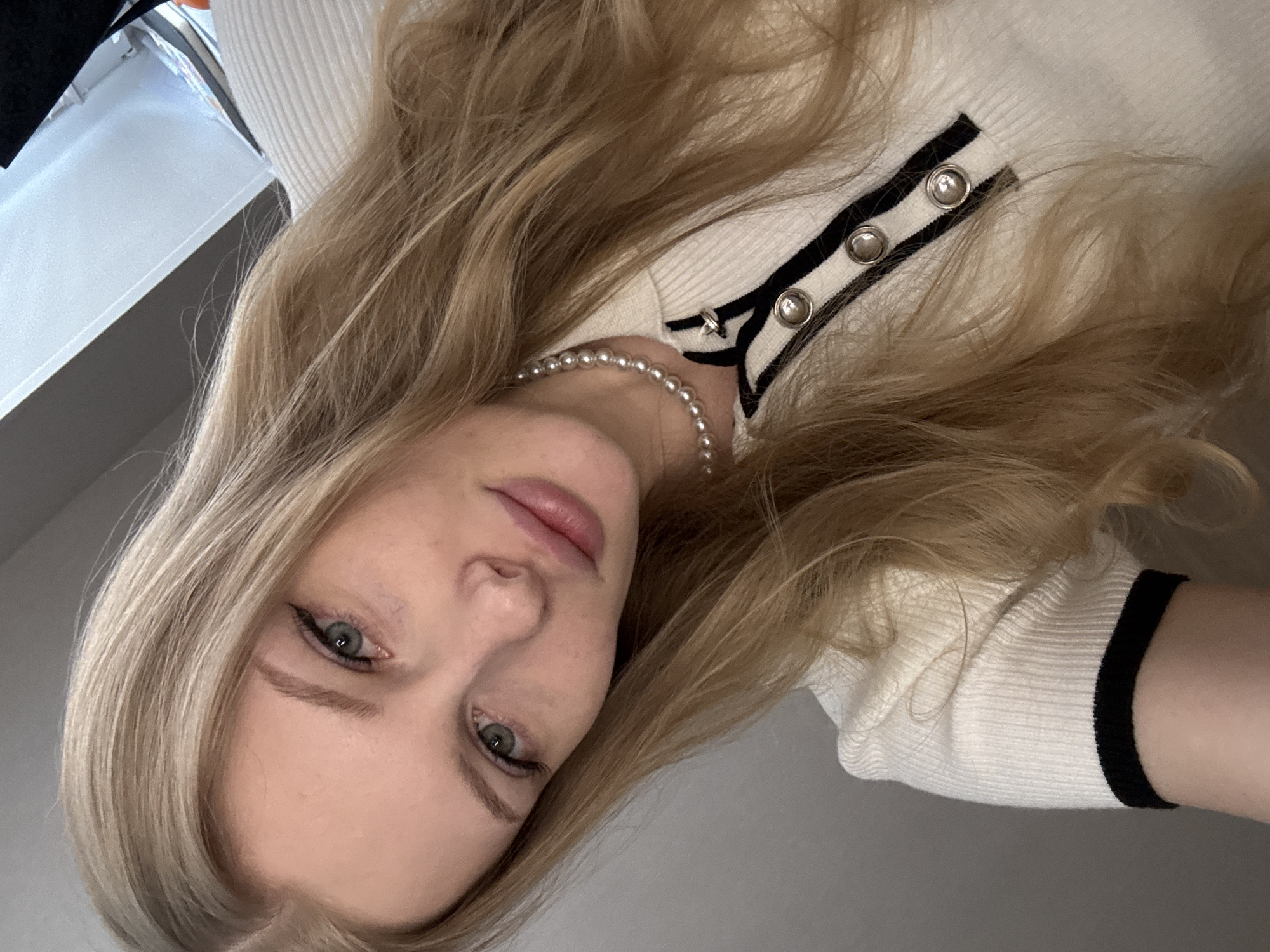 A young woman is depicted in a close-up, chest-up portrait. She has long, wavy blonde hair and fair skin, with visible freckles across her nose and cheeks. Her green eyes are accentuated with eyeliner, and her lips are full with a subtle pink tint. She is wearing a white, ribbed short-sleeved top featuring a black stripe along the collar and placket. The placket is adorned with three round, silver-toned buttons. A single-strand pearl necklace is visible around her neck. The background is out of focus, displaying indistinct light grey and white tones, indicative of an indoor setting. This scene is located in the city of Perm, Russia.