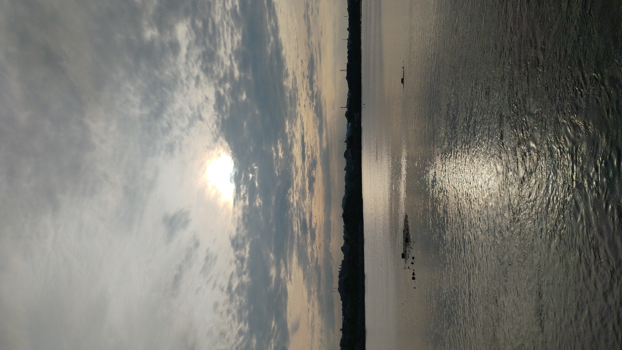 The image captures a serene, overcast day at the waterside, likely in Lhokseumawe, Indonesia, judging by the presence of a prominent body of water with a distant treeline. The sky is filled with layered, textured clouds in varying shades of grey, with the sun peeking through in the upper center, casting a bright, diffused light. Below the sky, a vast expanse of water dominates the lower half of the frame. The water's surface is rippled, reflecting the muted sunlight in a shimmering band that stretches from the horizon towards the viewer. In the foreground, a small cluster of dark rocks emerges from the water on the left side of the image, breaking the otherwise unbroken surface. The distant shoreline is a dark silhouette of rolling hills and scattered structures, including what appear to be communication towers, suggesting the proximity of a developed area. The overall atmosphere is tranquil and natural, with a sense of calm pervading the scene.
