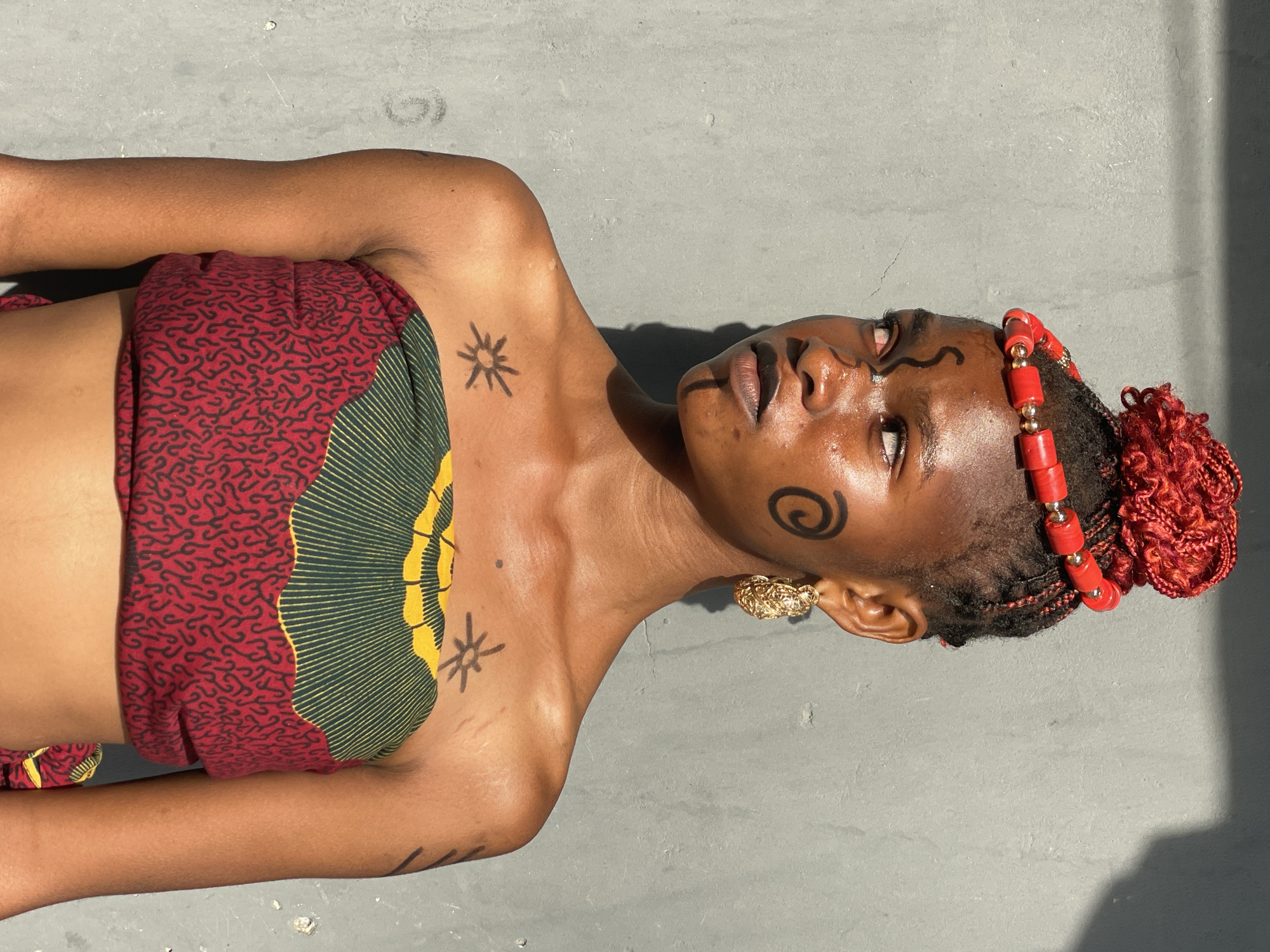 An eye-level, medium shot depicts a woman with dark skin lying on a grey surface, possibly concrete. She is wearing a strapless top composed of red fabric with a small black pattern, and a green fabric with a yellow sun-like pattern. She is adorned with tribal-style face paint in black ink on her forehead and cheek. Her hair is styled with a red bead headband, and her hair is partly black braids and partly bright red braided hair tied into a bun. A gold earring is visible. Tattoos in black ink are seen on her chest, shoulder, and arm. The scene is bathed in sunlight with a clear shadow indicating the direction of the light source. The location is not discernible, and there is no evidence indicating this is Badagry, Nigeria.