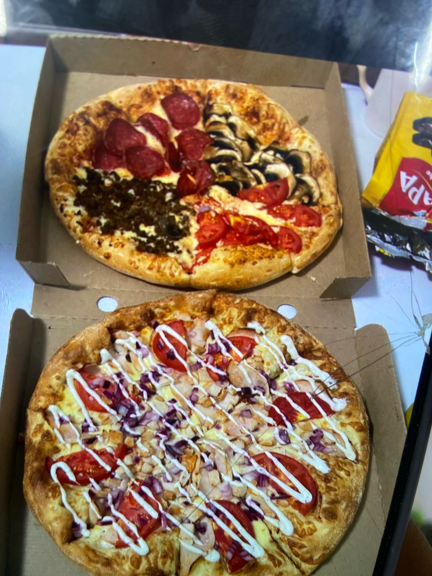 Two pizzas are shown in open pizza boxes. The top pizza is divided into four quadrants, featuring pepperoni, ground meat, mushrooms, and tomatoes. The bottom pizza is topped with chicken, tomatoes, red onions, and a white sauce drizzle. The pizzas appear to be in an indoor setting, possibly a home or a restaurant, with a yellow package visible in the background. The image does not provide any specific details to determine the location in the city of Aktau, Kazakhstan.