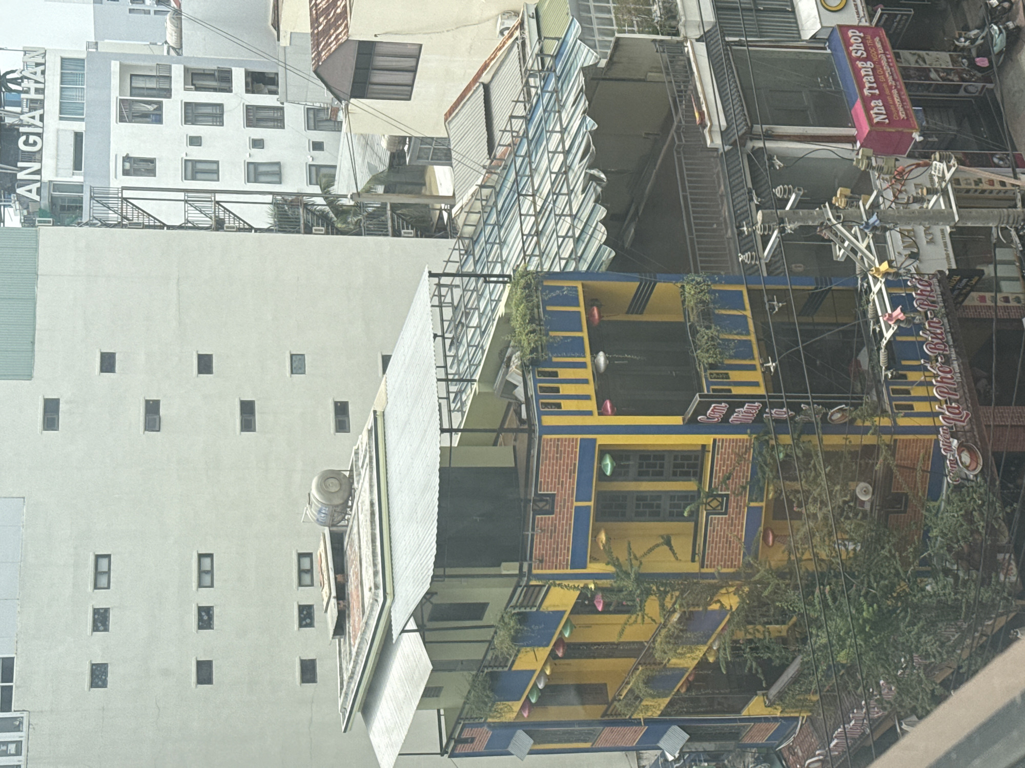 The image captures a street scene in Nha Trang Ward, Vietnam, exhibiting a mix of architectural styles and commercial activity. A tall, pale-colored building with rows of windows stands adjacent to a vibrant yellow and blue painted building, adorned with traditional lanterns and greenery. Adjacent is a building encased in a metal structure with a roof. Overhead utility wires crisscross the scene. Signage is visible for various businesses, including Nha Trang Shop and Pho Bun-Pho. The overall impression is of a bustling urban environment with a blend of residential and commercial buildings.