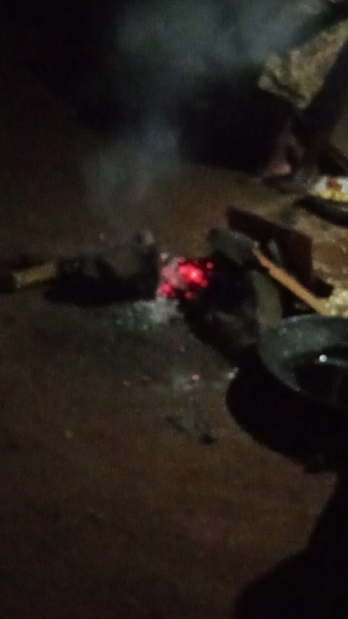 The low-light image captures a small, glowing fire or embers on a dark, possibly earthen, ground. The central focus is the bright red and orange glow of the burning materials, surrounded by scattered white ash. Smoke is visible rising from the fire, particularly on the left side of the frame.
The fire appears to be fueled by several dark, indistinct pieces of wood or charcoal. To the left of the embers, an elongated piece of wood or a similar object rests on the ground. To the right, a dark, round object, likely a pot or pan, is positioned close to the heat source, suggesting it is being used for cooking or heating. This object appears to be dark metal and has a slight reflective surface.
The setting is extremely dark and blurry, giving a sense of being indoors in a very dim room or outdoors at night. The background is mostly black, with faint, indistinguishable shapes that could be walls, other objects, or debris. The ground itself is dark and textured, resembling dirt or an unpaved floor. No people are visible in the scene, and there is no visible text. The overall atmosphere suggests a rustic, traditional cooking or heating setup, with fire as the primary light source.