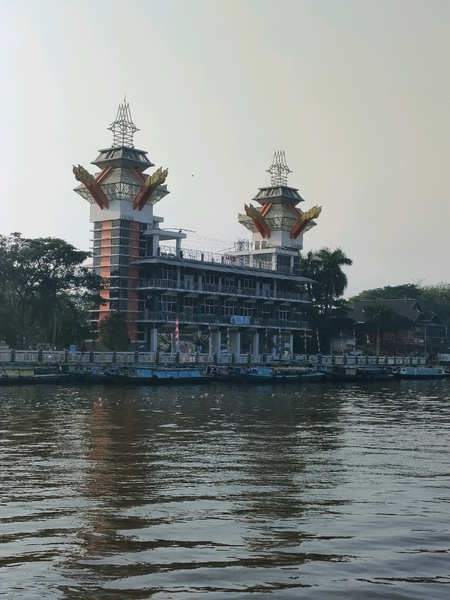 The image depicts a striking architectural structure located on the waterfront in Banjarmasin, Indonesia, likely along the Martapura River. The building features a distinctive, multi-tiered design with prominent, ornate roofs reminiscent of traditional Indonesian or Southeast Asian styles, possibly with avian or dragon motifs adorning the upper sections. These decorative elements are golden and appear to be stylized wings or crests. The main body of the building is a modern, multi-story structure with numerous windows, suggesting it could be a hotel, government building, or cultural center. Construction or renovation appears to be ongoing, with visible scaffolding and exposed framework on parts of the building.
In front of the building, a row of boats is moored along the riverbank, indicating this is a busy port or waterfront area. The boats are of various sizes and colors, primarily appearing to be utilitarian vessels, perhaps for transport or commerce. The river itself is wide and calm, with ripples on the surface reflecting the building and the sky. Trees and other smaller buildings are visible on the bank to the left and right of the main structure, suggesting a semi-urban or waterfront district. The sky is overcast or hazy, casting a soft, diffused light on the scene. No people are clearly discernible in the image, though the presence of the boats implies human activity in the vicinity. The overall impression is of a developing or active urban waterfront with a prominent, uniquely designed building as the focal point.