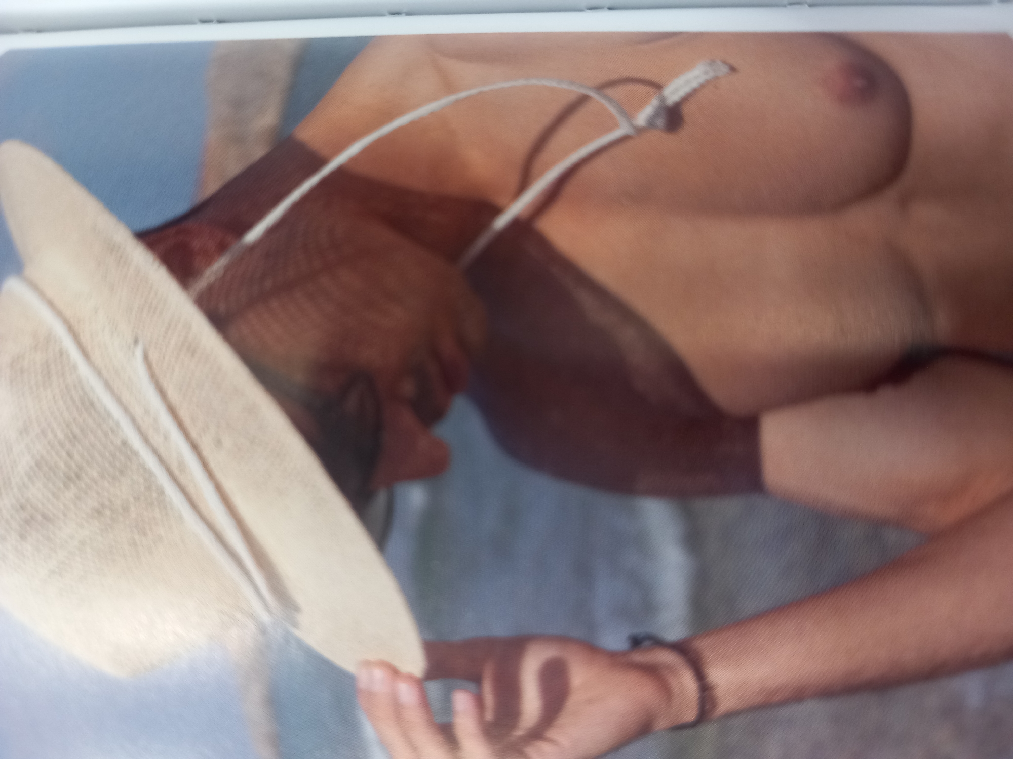 This media file shows a close-up, low-angle shot of a person's torso and arm, with a straw hat in the foreground. The person appears to be outdoors, possibly at a beach, with a blurred blue background suggesting water or sky. The person is partially undressed, with a string from the hat around their neck and a visible nipple. They are holding the hat, and their arm is resting on their body. The lighting suggests it might be daytime, and the overall mood is casual and relaxed. There is no visible text in the image.