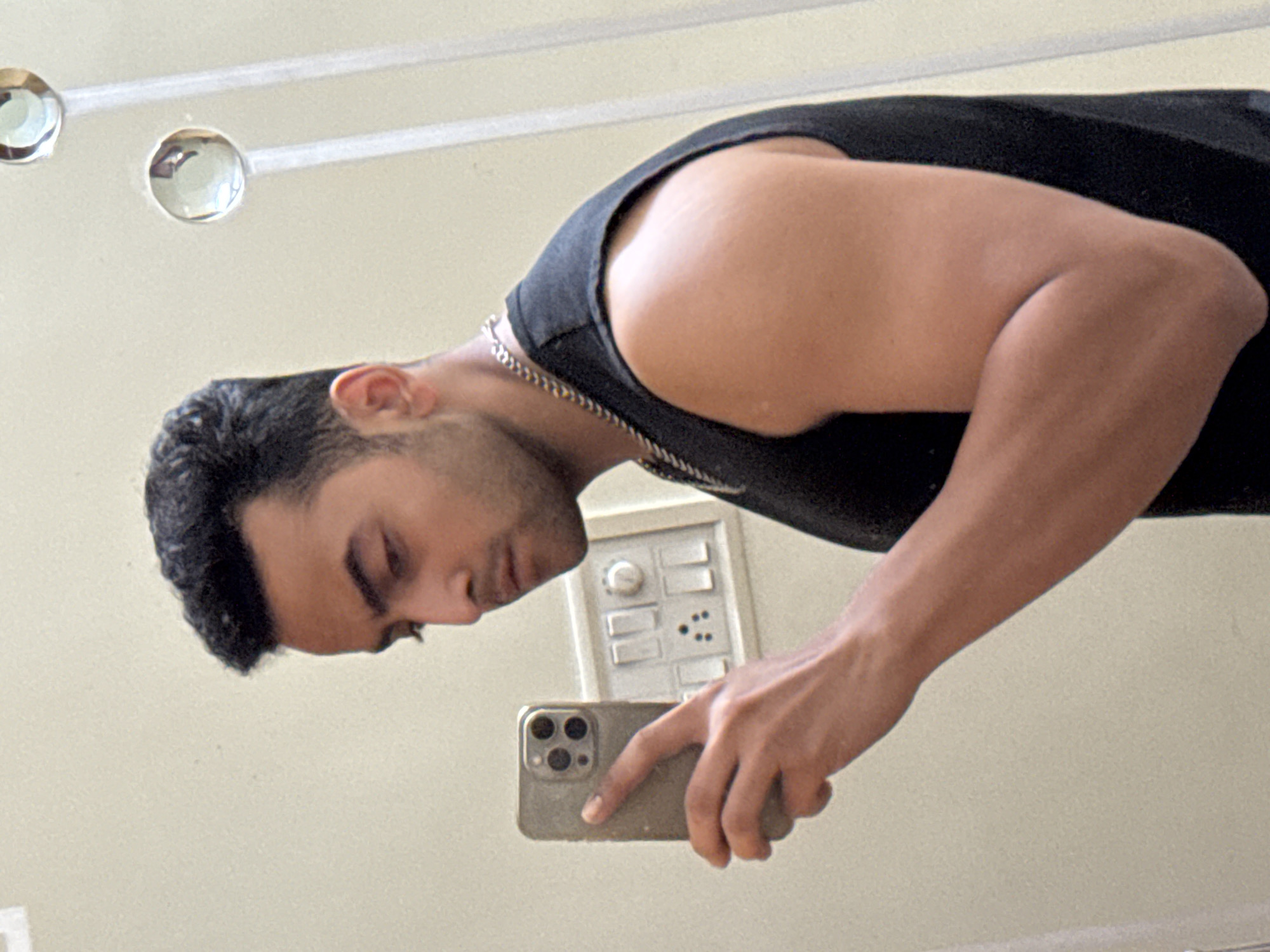 A man stands in front of a mirror, taking a selfie with a smartphone. He wears a black tank top and a silver chain necklace. A light switch panel is visible on the wall behind him. The location is not discernible as Kolkata, India based on the image.
