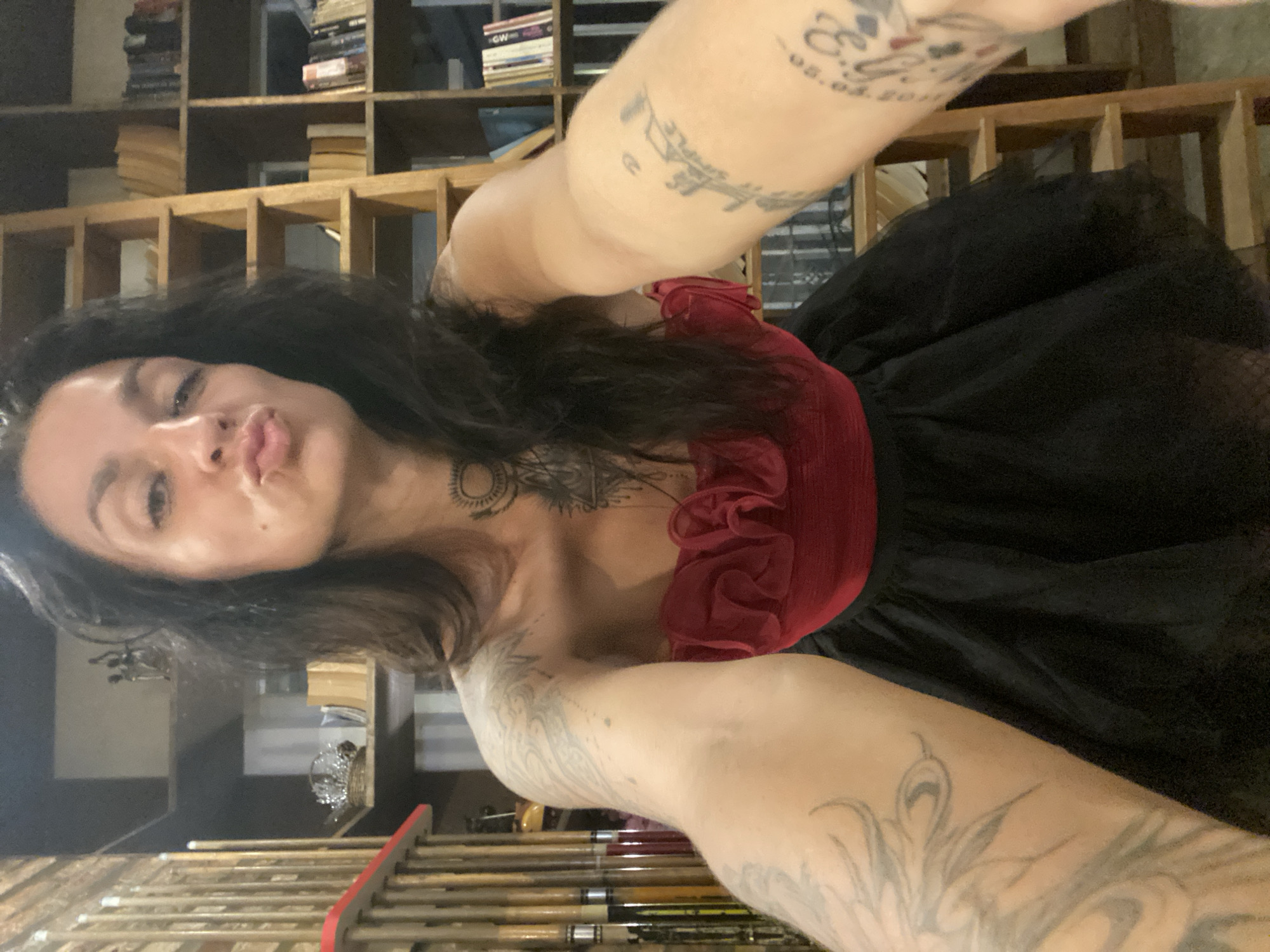 A woman with dark, long hair and multiple tattoos on her arms and torso, wearing a strapless red top with ruffled detail and a black tutu skirt, poses for a selfie. She is in an indoor setting that appears to be a room with bookshelves filled with books. A wooden ladder leans against the bookshelves. The environment does not suggest any specific location within Da Lat, Vietnam, as there are no discernible Vietnamese cultural or geographical markers.