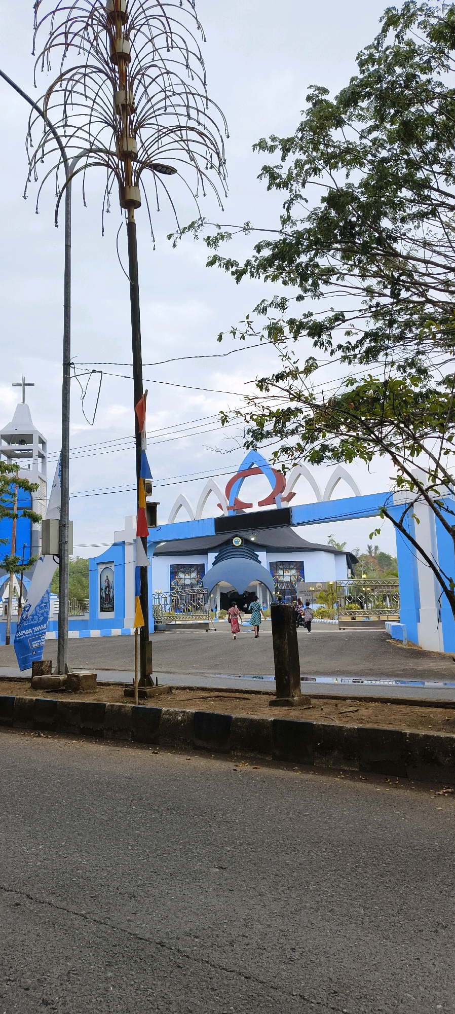 In the city of Waisai, Indonesia, a wide-angle view captures the entrance to a blue and white building, possibly a church or community center. The architecture features a prominent arched entryway adorned with a red and blue symbol, and white archways along the top. Flanking the entrance are large blue panels with white checkered patterns. To the left, a white bell tower with a cross is visible. In the foreground, a dark asphalt road runs across the bottom of the frame, bordered by a concrete curb. A tall, elaborate street lamp with a palm-tree-like structure stands to the left of the entrance, and a wooden pole with small flags is positioned near the center. Several people are walking towards and into the building, some dressed in colorful clothing. A few trees with green foliage are seen in the background and to the right of the frame, against a pale, overcast sky. The scene appears to be a public space on a cloudy day.