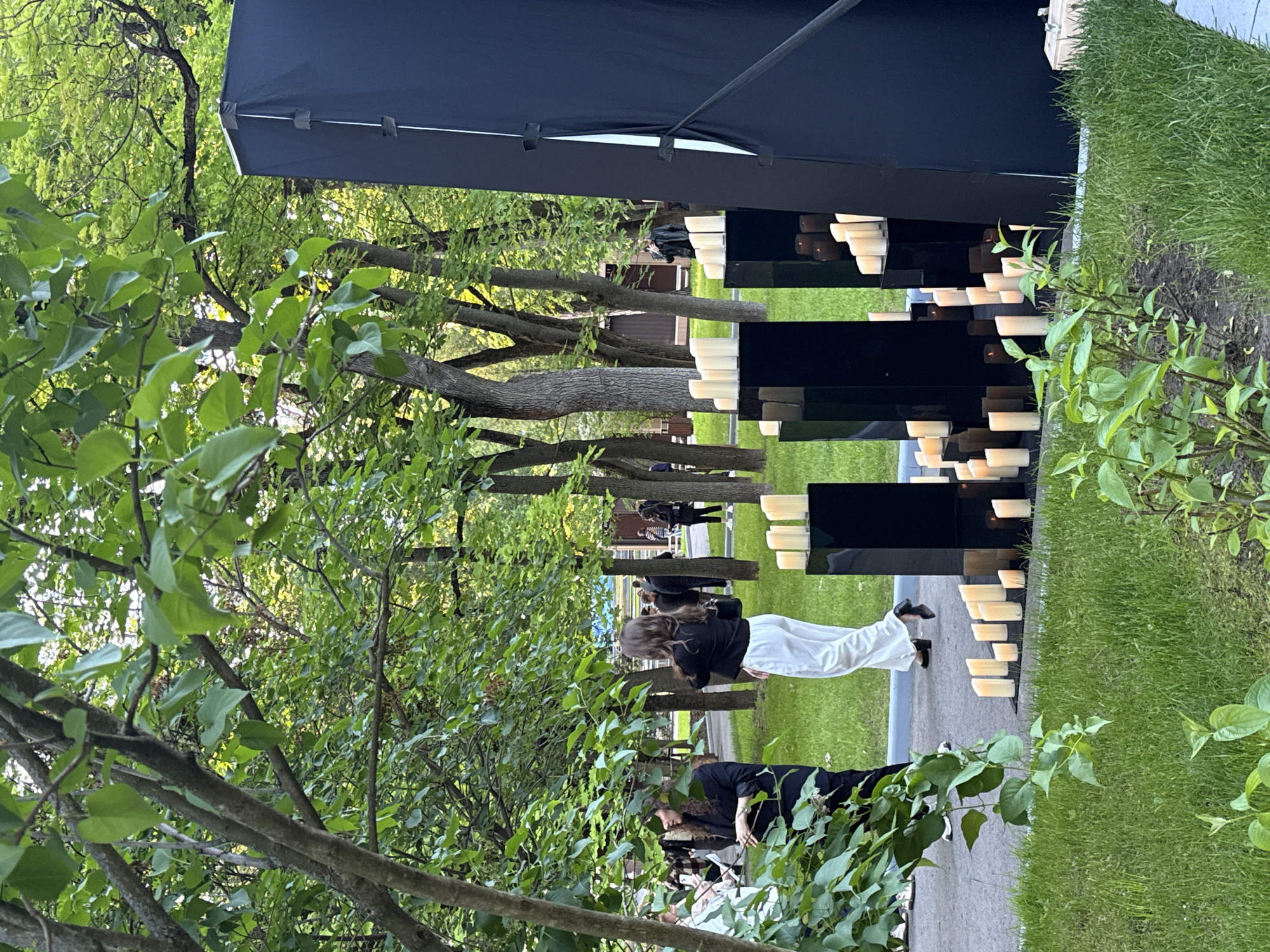 This image captures an outdoor scene, likely within a park or garden setting in Moscow, Russia. The environment is characterized by abundant green elements, including lush grass and numerous deciduous trees with vibrant green foliage, which frame the scene. A paved path or walkway traverses the area.
A significant feature of the scene is an artistic or commemorative installation. To the left, a large, dark-colored canopy or tent structure provides partial shade. Adjacent to the path, on the right side, an elaborate display is arranged. It consists of multiple black, rectangular plinths or pedestals of varying heights, creating a staggered, sculptural formation. Each plinth is adorned with several cream-colored pillar candles, contributing to a contemplative or ceremonial atmosphere.
Several individuals are present. In the midground, a woman with long, dark hair, dressed in a black top and white pants, is seen from behind, appearing to observe the candle display. Closer to the foreground, on the right, another person with dark, curly hair and a dark top is partially visible through the foliage. Other individuals are dispersed further back among the trees and along the path, suggesting a gathering or public event.
The time of day appears to be daytime, possibly late afternoon, under soft, natural lighting, indicative of pleasant, clear weather suitable for an outdoor gathering. The overall mood conveyed is one of tranquility and possibly reflection, stemming from the serene natural setting and the candle installation. No visible text is discernible in the image.