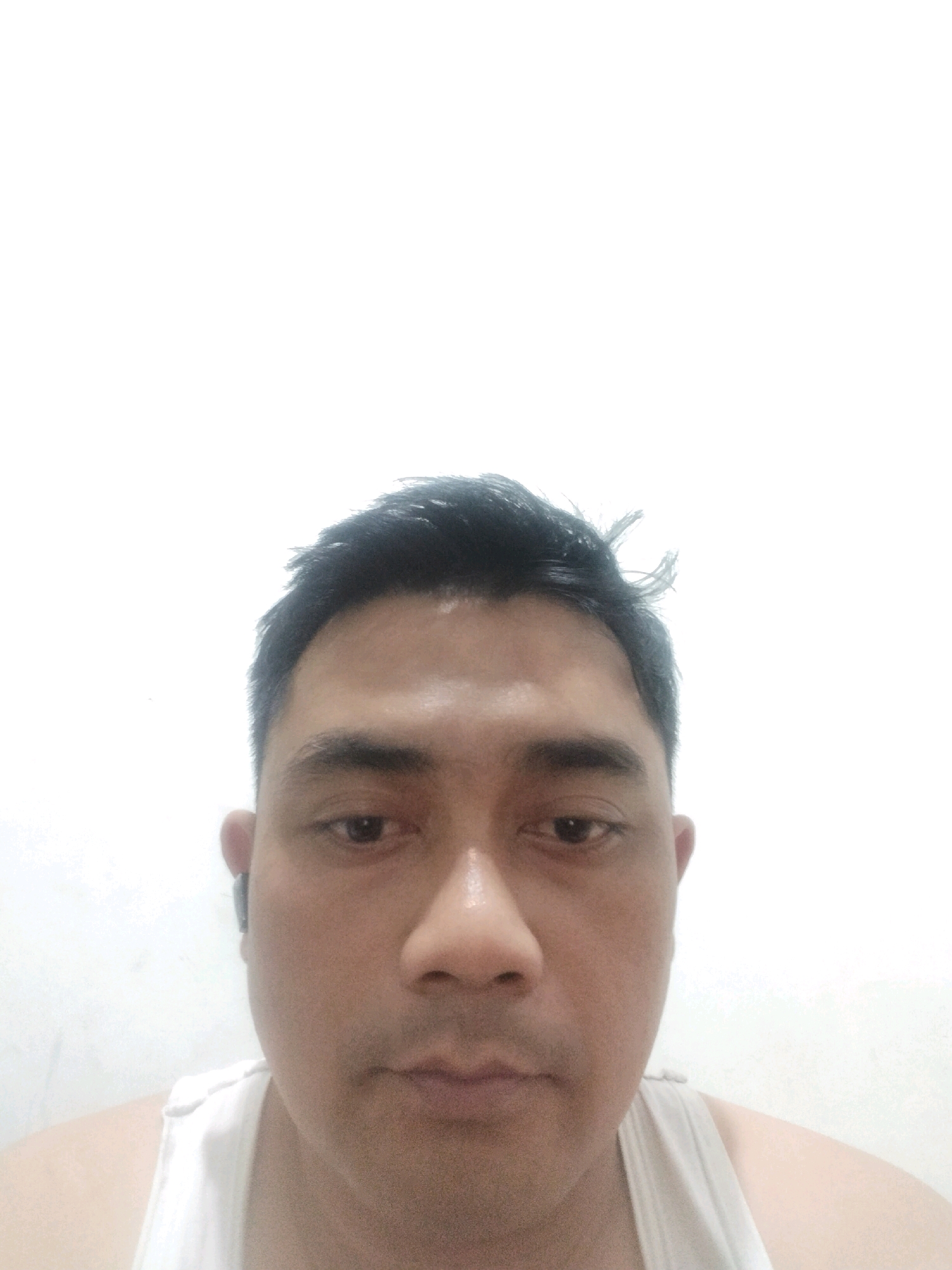 The image is a close-up, waist-up, front-facing photograph of a man in Malang, Indonesia. The man has dark hair, a fair complexion, and appears to be in his late 30s or early 40s. He is wearing a white tank top. He has a neutral expression and is looking directly at the camera. In his right ear, he is wearing a black wireless earbud. The background is a plain white wall, suggesting an indoor setting. The lighting is bright and even, with no shadows evident. The photograph is taken from a low angle, looking up at the man's face. The image is slightly out of focus, particularly around the edges.