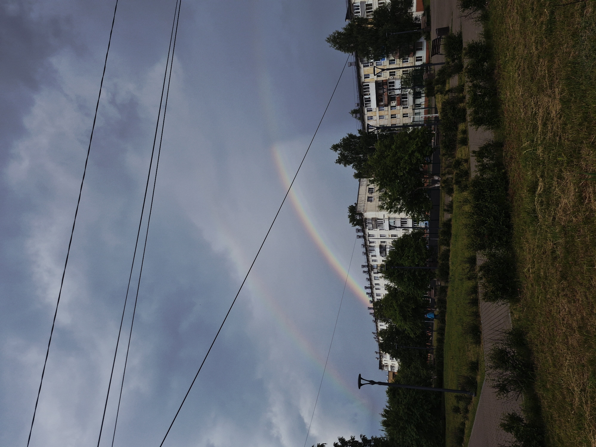 The image depicts a city park with apartment buildings in the background, under a dramatic, cloudy sky. Two distinct rainbows arc across the sky, with the fainter one appearing above the more vibrant one. In the foreground, a paved walkway and green grass suggest a recreational area. Several trees are visible, some taller and some shorter bushes, along with streetlights and park benches. The apartment buildings are typical of Soviet-era architecture, with numerous windows and balconies. The overall atmosphere is subdued due to the overcast conditions, but the presence of the double rainbow adds a touch of natural wonder to the urban landscape. The image appears to be taken in Chaikovsky, Russia, given the architectural style and the specific detail of the double rainbow.