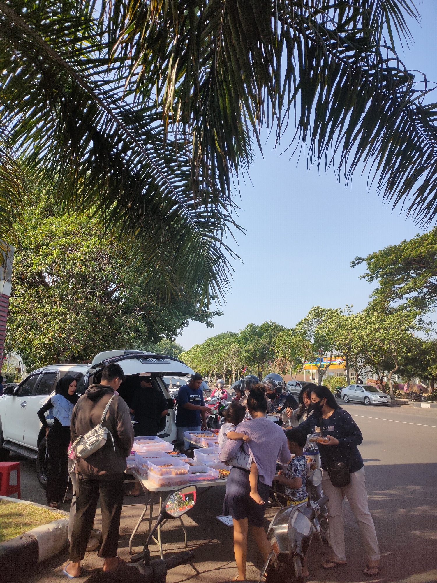 In Ngasem, Indonesia, a street scene unfolds under the dappled sunlight filtering through palm fronds. A white SUV with its trunk open serves as a backdrop for a makeshift outdoor market. A table laden with clear plastic containers filled with various food items is set up on the asphalt. People are gathered around the table, some examining the food, others in conversation. A woman in a light blue headscarf and dark pants stands near the SUV, her hand on her hip, looking towards the table. A man wearing a gray hoodie and carrying a bag slung over his shoulder stands in front of the table, facing away from the camera. Another man in a dark blue shirt is seen behind the table, interacting with a customer. To the right, a woman in a dark shirt and light-colored pants holds a food container, engaged in a transaction. A young child is held by a woman in a purple top, and a boy in a patterned shirt stands nearby, all facing the table. Motorbikes are parked on the side, suggesting a busy street. The environment includes lush green trees lining the road, a clear blue sky, and other vehicles in the background, indicating a typical urban setting.