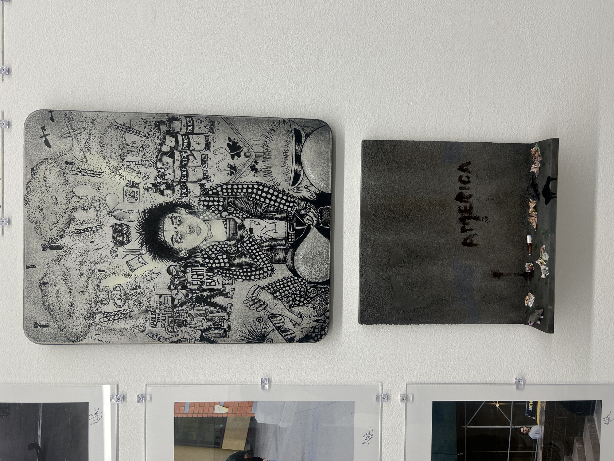 The image displays an art installation featuring two main pieces mounted on a white wall, with parts of other artworks visible below.
The dominant piece on the left is a large, rectangular, monochrome illustration with rounded corners. Rendered in a dense, graphic, and somewhat chaotic style, it depicts a central figure with spiky hair and a dotted shirt, looking forward. Surrounding this figure is a complex scene filled with symbols of protest and conflict. To the right of the central figure, a line of riot police is visible, with the word "POLICE" repeatedly written on their shields. Below them, protesters hold signs, one clearly stating "ABOLISH THE POLICE," another appearing to say "FIGHT BACK," and a third "NO WAR." Other elements in the illustration include falling bombs, a mushroom cloud, an airplane, birds, keys, a bottle, and a balloon with the text "YOUR BAND." The background also contains the word "CAUTION."
To the right of the illustrated piece is a smaller, dark, rectangular object, possibly made of metal or a similar textured material. The word "AMERICA" is written across its surface in a dark, possibly etched or painted, script. The bottom right corner of this piece is bent forward, forming a small shelf on which various small pieces of debris, including what appear to be bottle caps, crumpled paper, and a miniature rat figure, are affixed.
Below these two main artworks, the tops of three framed prints or photographs are visible, held by clear plastic clips. One of these lower prints shows a partial street scene with scaffolding, and the word "NEW" is faintly visible.
The overall scene presents a multi-media art display with themes that appear to be critical or commentary on society, policing, and the state of America.
