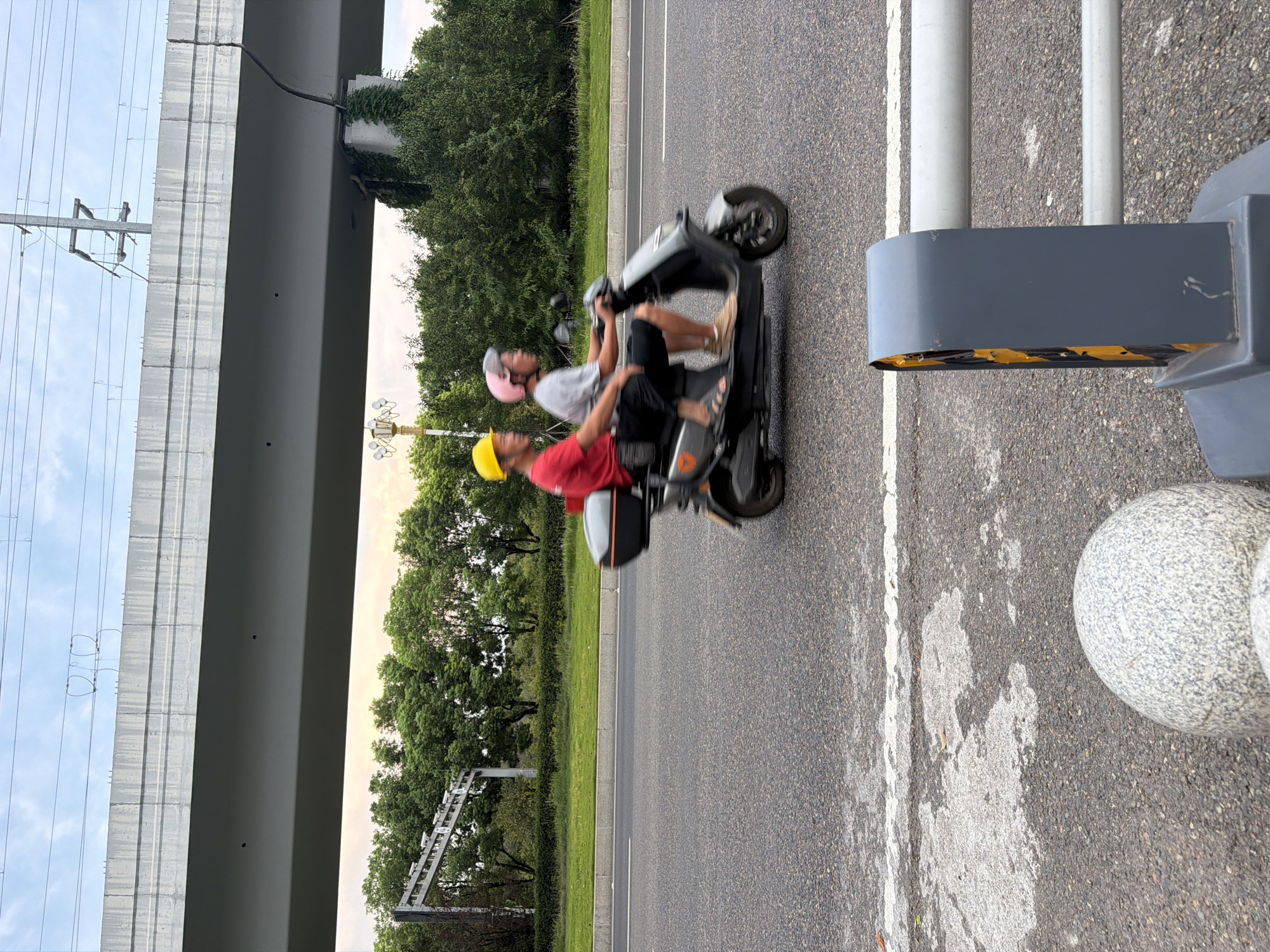 In Jiangjin, China, two individuals are riding on a black electric scooter on a paved road. The person in the front is wearing a yellow helmet and a red shirt, while the person in the back is wearing a pink helmet and a grey t-shirt. They are both wearing casual clothing. The scooter has a black storage box on the back. The road has a white line marking its edge. In the background, there is lush green foliage and trees. Above the trees, a concrete overpass or bridge is visible, supported by concrete pillars. Power lines are strung across the sky above the overpass. The sky is partly cloudy with a light sky. In the foreground, a gray, cylindrical concrete bollard is visible on the right side of the image, and a portion of a gray metal barrier is also present. The overall scene depicts everyday urban transportation.
