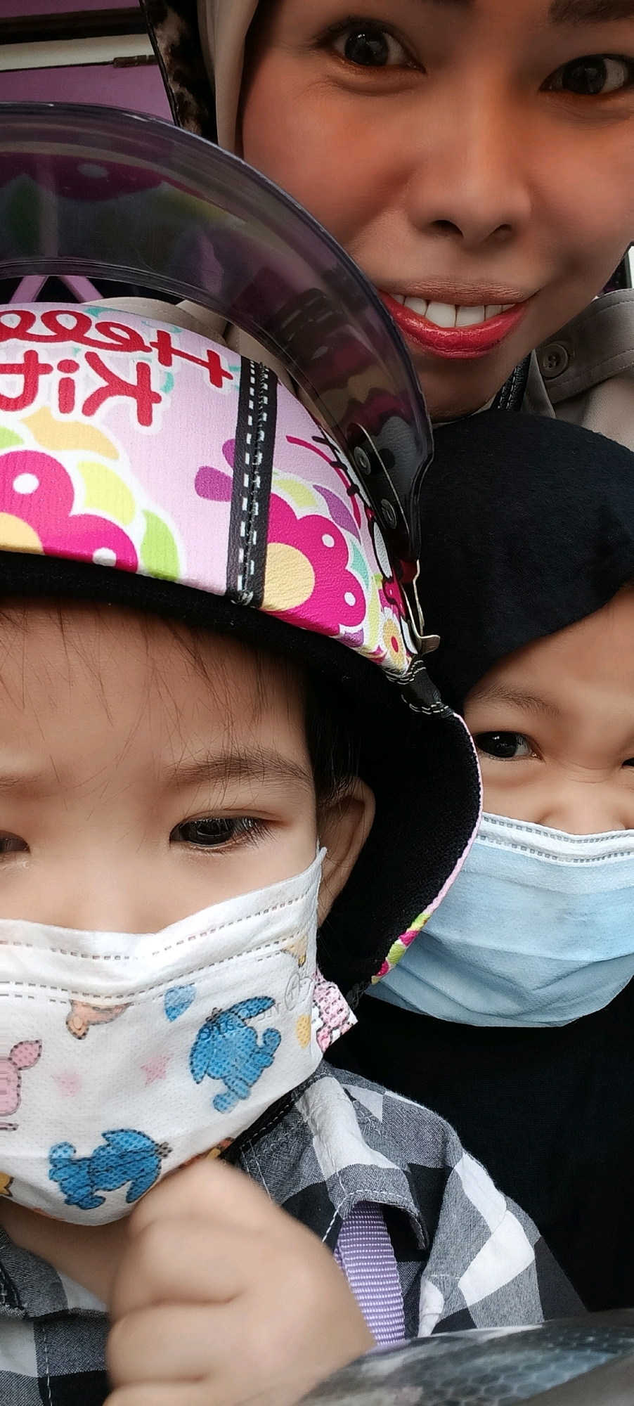 A close-up, head-and-shoulders portrait shows three individuals wearing face masks, suggesting a public health context. The person in the foreground, on the left, is a child with dark hair, wearing a white mask decorated with blue cartoon characters. Their head is partially obscured by a colorful helmet featuring a pink and white design with floral motifs and the text "Kiki." To the right of this child is another individual, possibly a younger child, also wearing a mask, this one light blue. Their eyes are visible, and they appear to be looking towards the camera. In the background, an adult, likely a woman, is visible. She is wearing a hijab and a light-colored jacket. Her eyes are open, and she is smiling, revealing red lipstick. The overall lighting suggests an outdoor setting, possibly during the day. The image appears to have been taken in Kuala Kapuas, Indonesia.