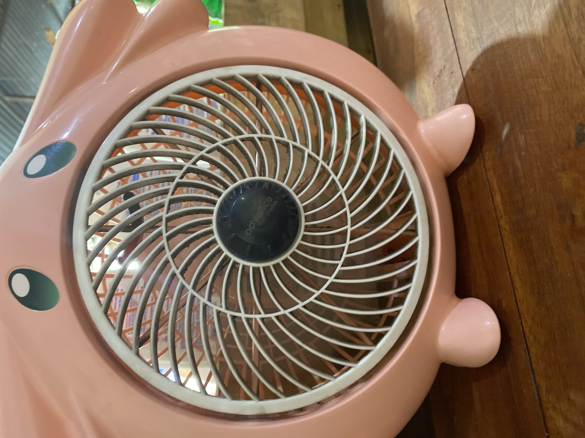 The image shows a close-up of a pink fan designed to resemble a cartoon character, possibly Jigglypuff from Pokemon. The fan is sitting on a wooden surface, likely a table or floor, in what appears to be an indoor setting with a corrugated metal roof visible in the background. The fan has large, round, green eyes with white pupils, and its body is bulbous. The fan's grill is white with orange blades visible behind it. The setting suggests a casual environment, possibly a home or a small shop in Ta', Indonesia. There are no people visible in the image.