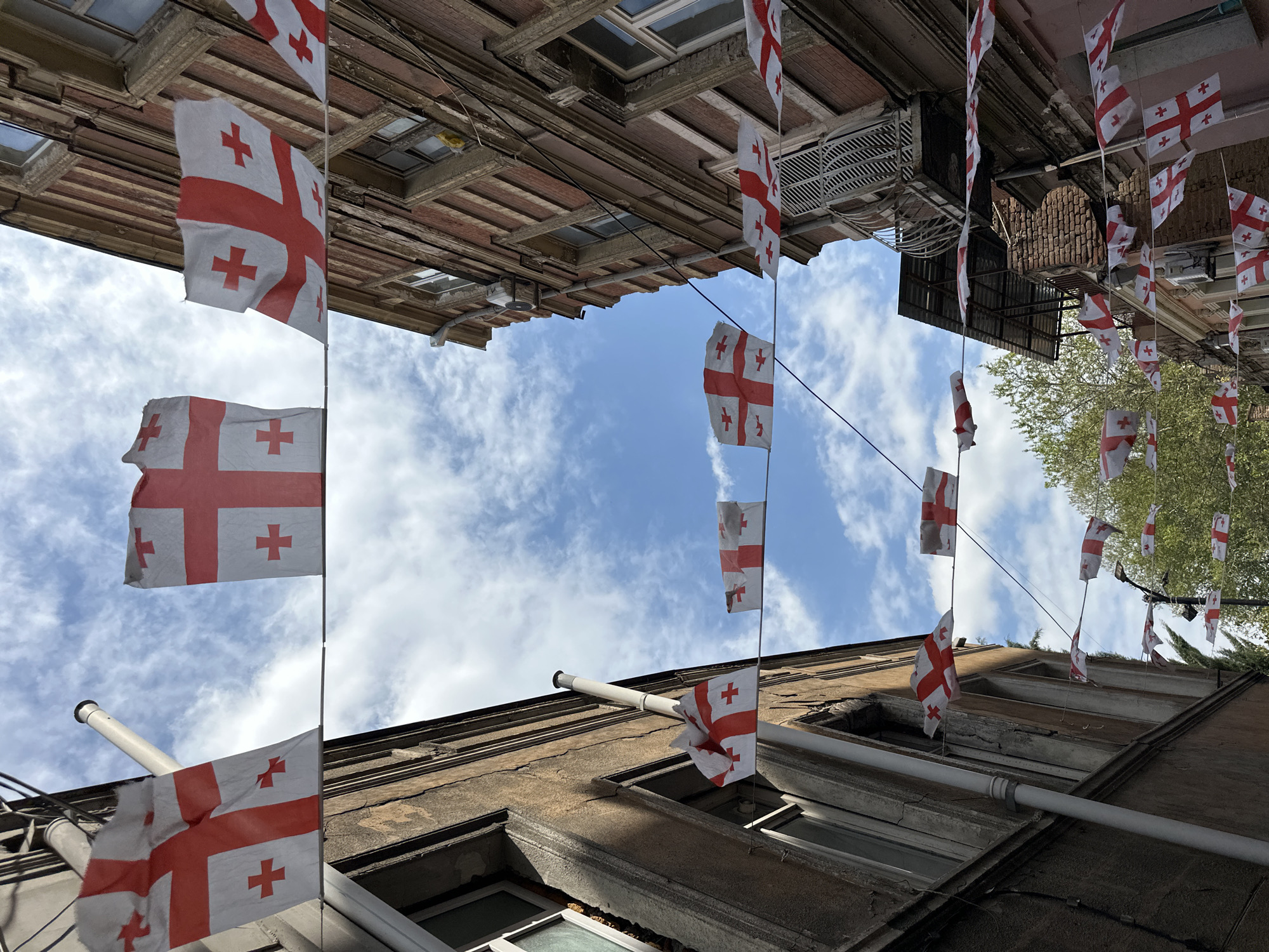 The image, oriented correctly, captures a low-angle view looking up a narrow passage between old buildings in Tbilisi, Georgia. Numerous Georgian flags are strung on wires overhead, creating a decorative canopy against a bright blue sky dotted with scattered white clouds.
The flags are predominantly white, each featuring a large red St. George's cross extending to the edges, and four smaller red Bolnisi crosses in the quadrants. Many flags appear slightly worn and are gently ruffled by the wind. They are arranged in multiple rows, crisscrossing the space between the structures.
On both sides, the weathered facades of multi-story buildings rise, showcasing an older architectural style. The buildings have textured, possibly stucco or stone exteriors with visible cracks and details around windows. Some window frames are dark, and exposed pipes or conduits run along the walls. Green foliage, likely from trees, can be seen extending above the rooftops on the right side of the frame.
The clear sky and bright illumination indicate it is daytime, suggesting good weather. The presence of the Georgian national flags, combined with the historical appearance of the buildings, firmly places the scene in a patriotic or celebratory context within Tbilisi. The visible symbols are the prominent red crosses on the flags.