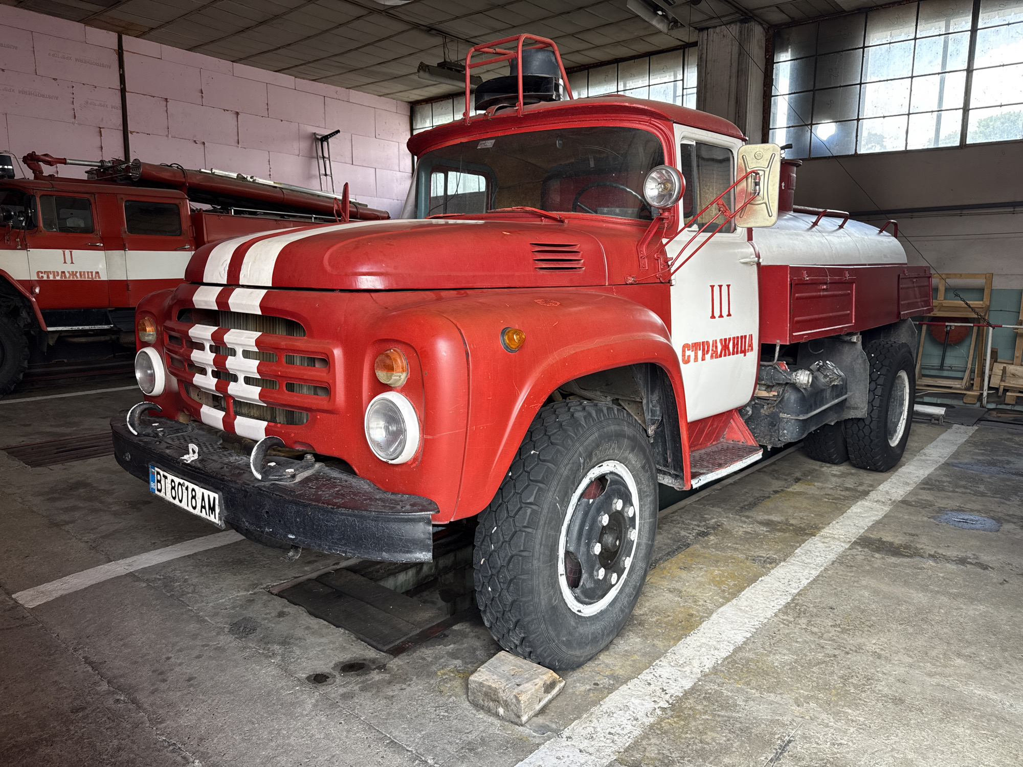 The image shows two red and white fire trucks parked inside a garage or large building. The truck in the foreground features the number "III" and the Cyrillic word "СТРАЖИЦА", which is the name of the city of Strazhitsa in Bulgaria. A number plate reading "BT 8018 AM" is visible on the front bumper. The truck is parked with its front tire resting on a piece of wood. Another truck can be seen in the background with the number "II" and the same Cyrillic inscription. The garage has striped parking lines.