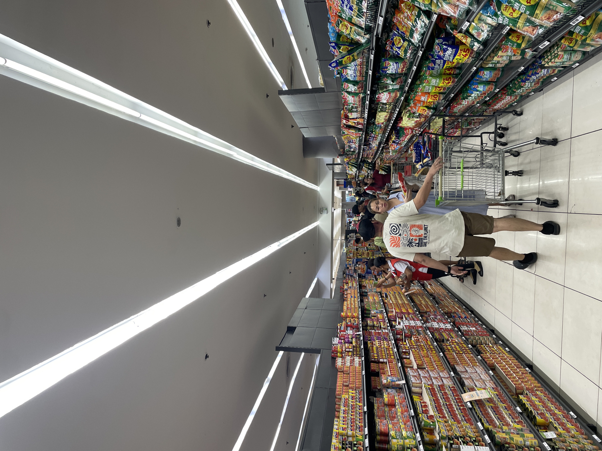 In a supermarket aisle in Davao City, Philippines, shoppers navigate between shelves stocked with canned goods and packaged snacks under bright, elongated fluorescent lights. A man in a light-colored t-shirt and shorts is pushing a shopping cart, facing away from the camera. To his left, a woman in a blue dress holds a product. Further down the aisle, other shoppers are visible, including a store employee in a red vest. The shelves are filled with various food items, organized neatly, and the floor is tiled in a light color. The scene captures a typical retail environment with people engaged in grocery shopping.