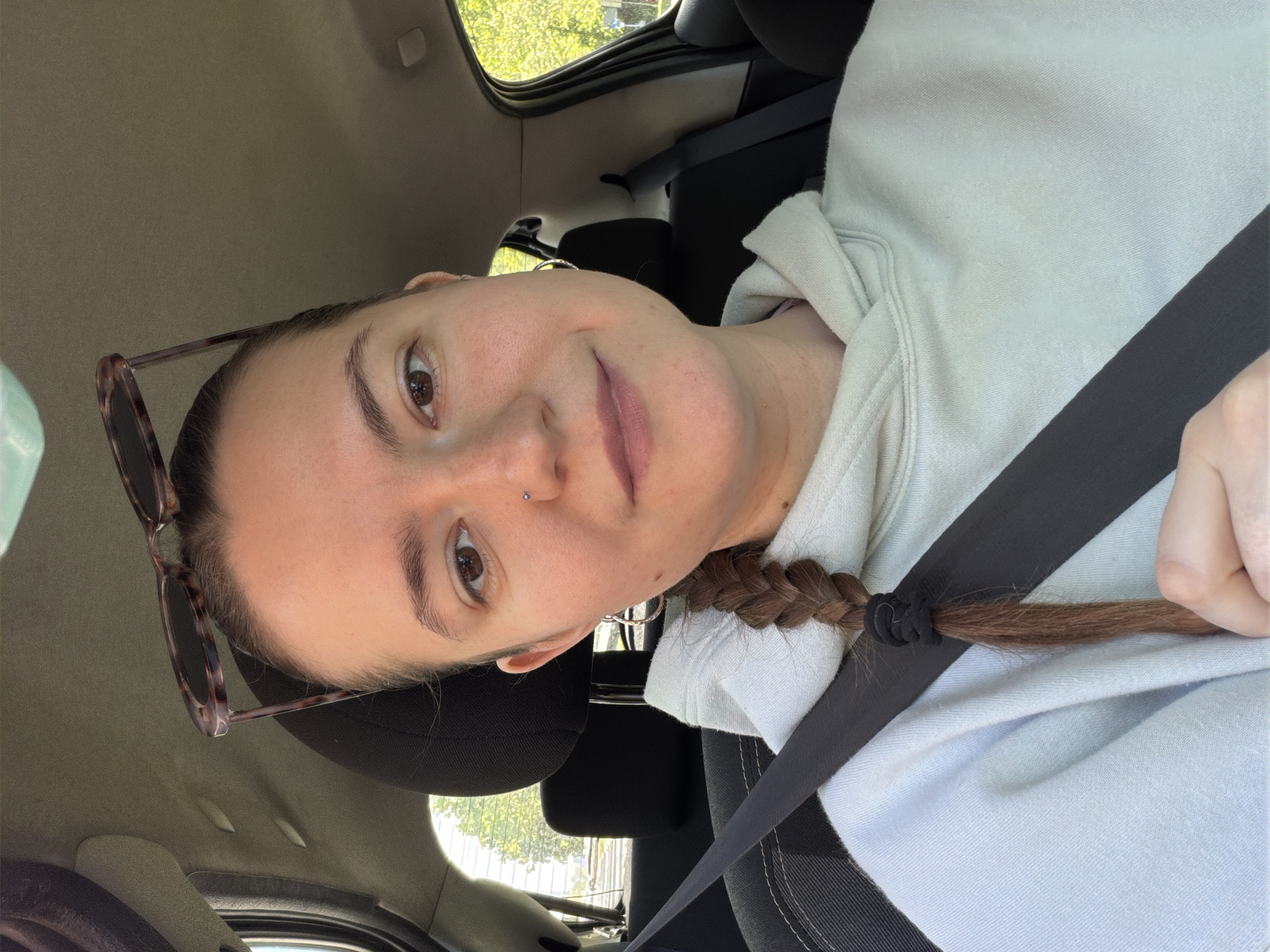 This image captures a selfie of a young woman sitting in the passenger seat of a car, likely in Belgrade, Serbia. The woman, with fair skin and dark brown eyes, gazes directly at the camera with a neutral to slightly soft expression. She has reddish-brown hair, styled in a single braid secured with a black elastic, falling over her left shoulder. A small silver stud is visible on her nose piercing, and she wears a simple hoop earring on her visible ear. Sunglasses with tortoiseshell frames are perched on top of her head.
She is wearing a light blue or off-white hooded sweatshirt. A dark seatbelt crosses diagonally over her chest and lap, indicating she is buckled in. The interior of the car is visible, featuring dark gray or black seats and a light-colored headliner. Through the car windows, green foliage from trees and glimpses of what appears to be an urban or suburban outdoor setting are visible, bathed in natural daylight. The lighting suggests it is daytime and potentially sunny. No text is visible in the image.