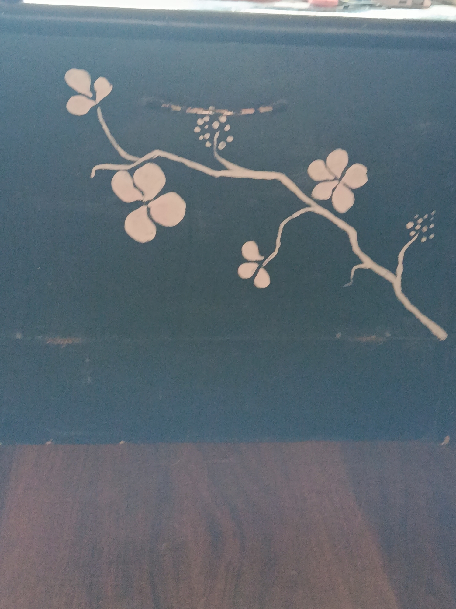 The image shows a close-up view of a dark blue surface with a white floral design. The design depicts a branch with several flowers and buds. The flowers have multiple rounded petals, and the buds are clustered together. The branch is thin and winding, with smaller twigs extending from it. The background is uniformly dark blue, with some slight texture visible. The bottom portion of the image shows a blurred area of brown, which appears to be a wooden surface. There are no people or actions depicted in the image. The location in the city of Dachne, Ukraine is not discernible from the image itself.