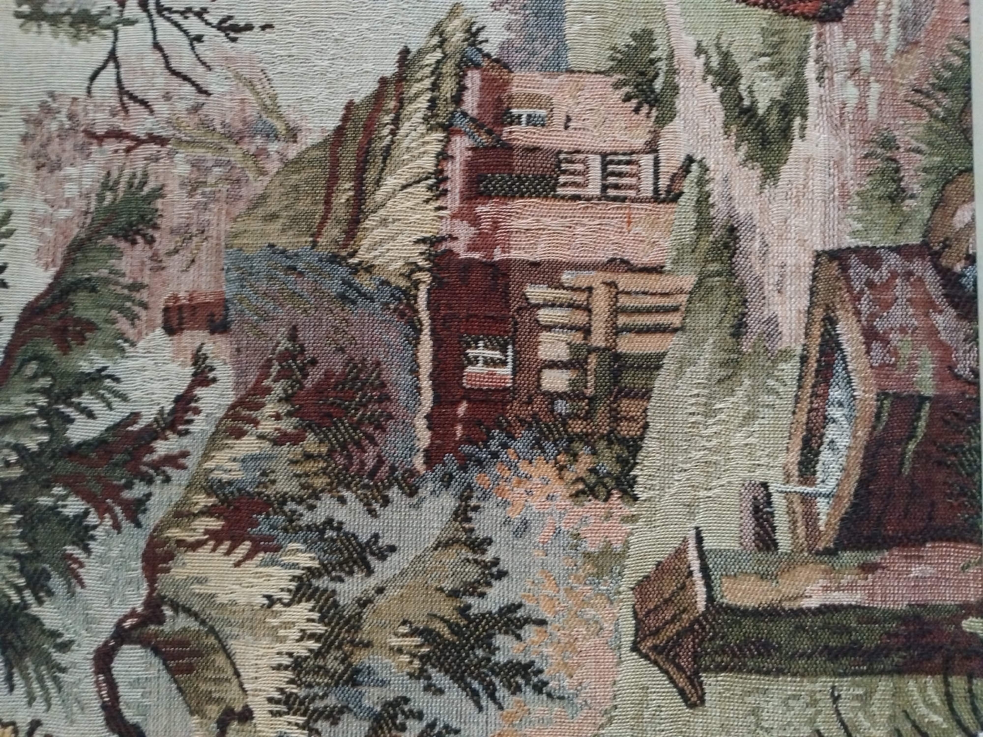 The image is a tapestry depicting a rustic scene. In the foreground, a stone well structure with a wooden roof is visible. Behind it, a path leads towards a quaint, thatched-roof cottage with red walls and windows. The cottage is nestled amongst lush greenery and tall trees, with branches extending over the scene. The overall impression is one of a tranquil, rural setting. The tapestry's texture suggests it is made of woven threads, creating a detailed and textured visual. There is no information in the image that relates it to Akhaltsikhe, Georgia.