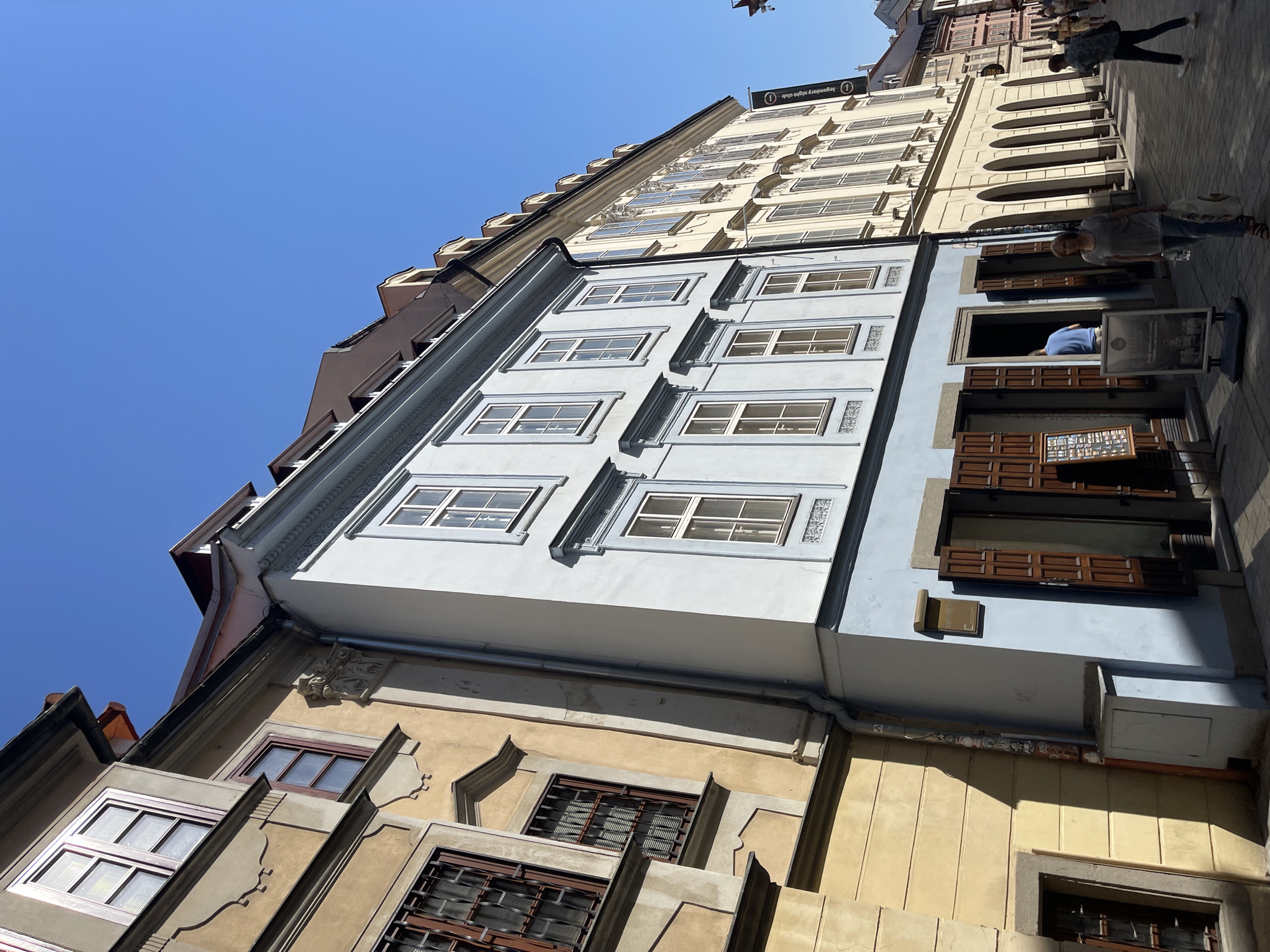 The image shows a street scene in Bratislava, Slovakia, with a narrow cobblestone street flanked by historical buildings. The buildings are multi-story with varied architectural styles and colors, including yellow, light blue, and cream. The facades feature multiple windows, some with ornate frames and shutters. The sky is clear and blue. On the right side of the street, there is a banner hanging from a building that reads "legendary night club". Several people are visible walking along the street. One woman is walking towards the camera carrying a bag, and another person is walking away from the camera in the background. A man in a blue shirt is visible standing inside one of the buildings. There are signs and displays outside some of the establishments. The scene captures a sunny day in a European city.