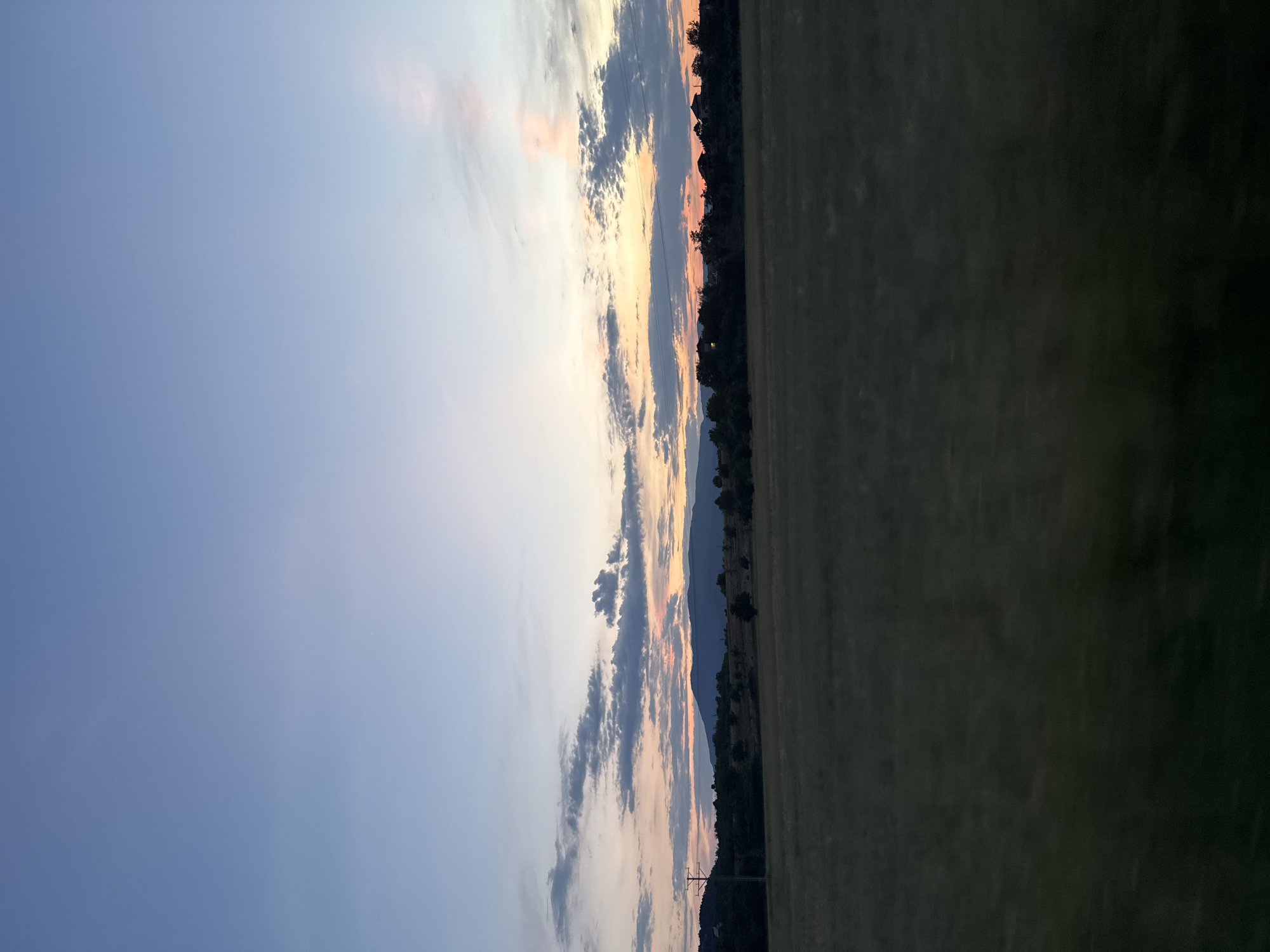 A wide-angle, low-angle shot captures a dusk scene over a field in Slokoshtitsa, Bulgaria. The sky transitions from a pale blue at the top to a warm blend of pink, orange, and yellow near the horizon. Scattered, wispy clouds are present in the mid-sky. A dark, undulating mountain range forms the backdrop, silhouetted against the colorful sky. In the middle ground, a line of trees with some houses nestled within them runs horizontally across the frame. To the left, a solitary utility pole with wires stretches upwards. The foreground is dominated by a dark, out-of-focus field, suggesting the photograph was taken from a moving vehicle.