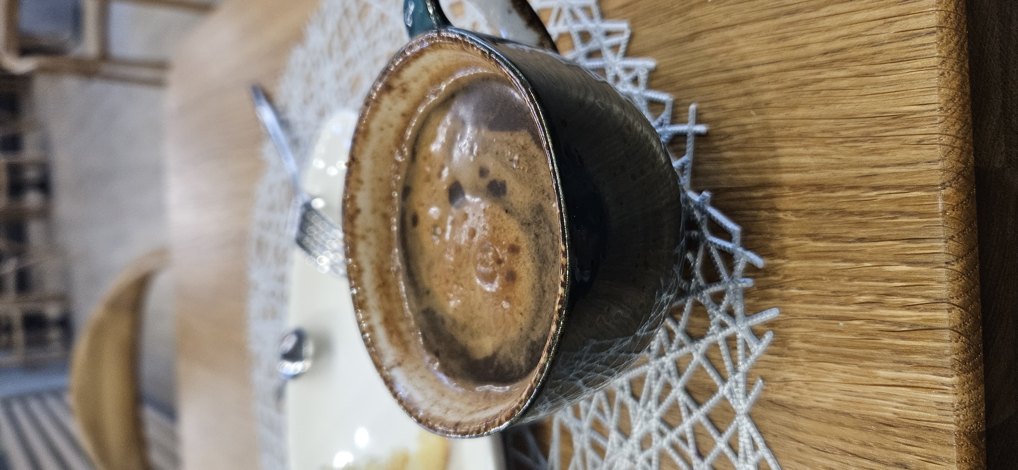 A close-up, vertical shot shows a dark, textured mug filled with frothy coffee sitting on a light-colored woven placemat on a wooden table. The coffee has a rich brown color with lighter swirls on the surface. The mug has a dark exterior with a lighter, textured rim. In the background, out of focus, a white plate with silverware is visible. The setting appears to be a cafe or restaurant, possibly in Vitsebsk, Belarus, judging by the style of furniture and general ambiance. The lighting is soft and natural, suggesting an indoor setting during the day.