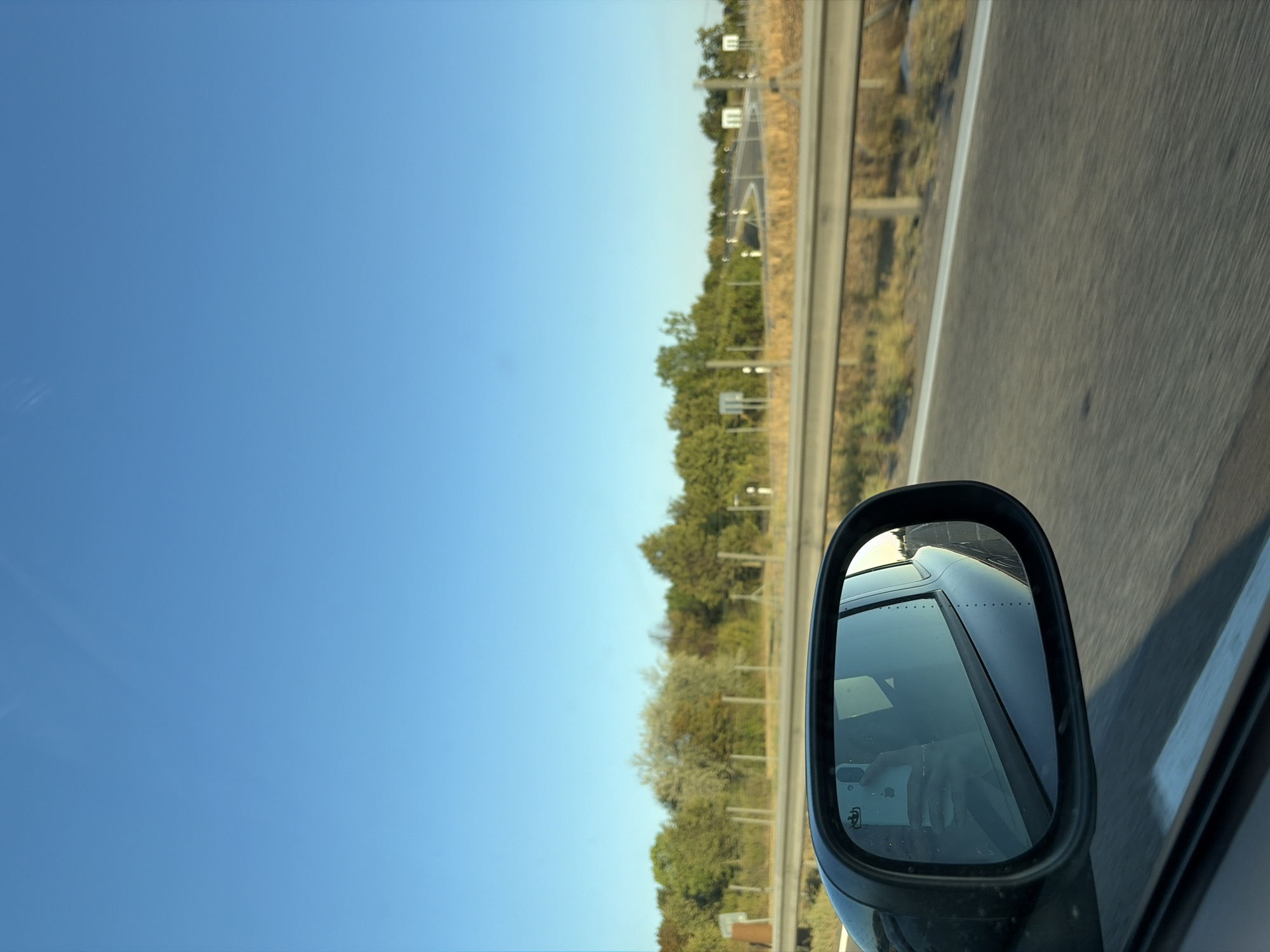 The image depicts a view from a moving vehicle on a sunny day, likely on a highway outside the city of Šintava, Slovakia. The foreground features a side-view mirror, reflecting the interior of the car, including a person holding a smartphone. The road surface is visible, with motion blur indicating speed. Beyond the road, a metallic guardrail runs parallel, separating the highway from a grassy median and a tree line. The trees exhibit varied shades of green and some hints of autumn colors. In the distance, the road curves, and white road markers are visible. The sky is a clear, bright blue, with a few faint wisps of clouds. The overall impression is one of travel on a clear day, with a focus on the driving experience and the surrounding landscape.