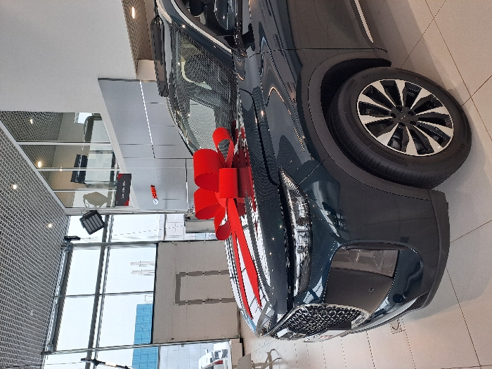 A dark-colored vehicle, likely a car or SUV, is adorned with a large red bow. The car is parked indoors, presumably in a showroom, given the tile floor and glass windows. The vehicle's front is prominently displayed, including the wheel and headlight. The background features a grid pattern, possibly part of the building's architecture, and exterior views through the windows.
