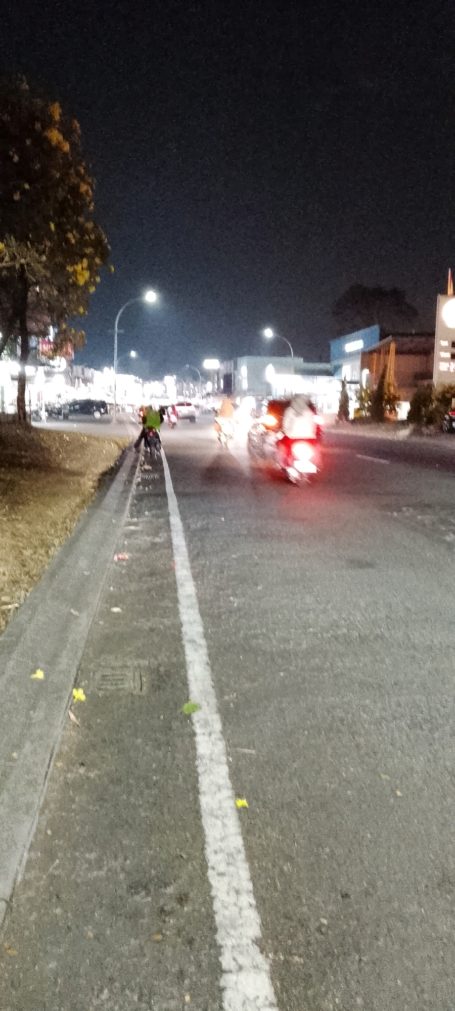 This is a nighttime photograph of a street in Cileungsi, Indonesia. A motorcycle is visible in the foreground on the left side of the image, with its rider wearing a green vest. The motorcycle is positioned on the edge of the road, near a curb and drainage grate. The road is asphalt and has a white line marking the lane. Further down the road, other vehicles, including motorcycles and cars, are present, some with their lights on, creating streaks of light due to the long exposure. The background features buildings illuminated by streetlights and shop signs, indicating an urban or commercial area. A tree with yellow leaves is on the left side of the image. The overall atmosphere is dark, with artificial lights illuminating the scene.