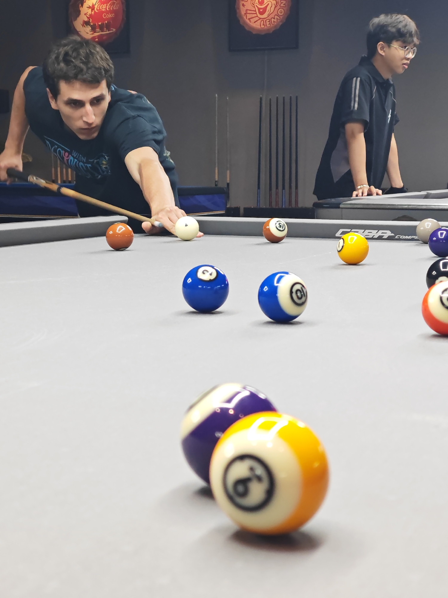 Two young men are playing pool in a dimly lit room. The man in the foreground, with dark curly hair, is leaning over the table, holding a cue stick and preparing to strike a white ball. He is wearing a dark blue t-shirt with a graphic design. To his right, a white ball and a solid orange ball are visible on the grey felt surface of the pool table. In the background, another young man with short dark hair and glasses, wearing a black polo shirt with grey accents, is leaning on the table. Behind him, several pool cues are neatly arranged on a rack. The environment appears to be an indoor pool hall with posters on the wall, including one that resembles a Coca-Cola advertisement. The scene is set in Đà Nẵng, Vietnam, likely at a recreational facility or entertainment venue.