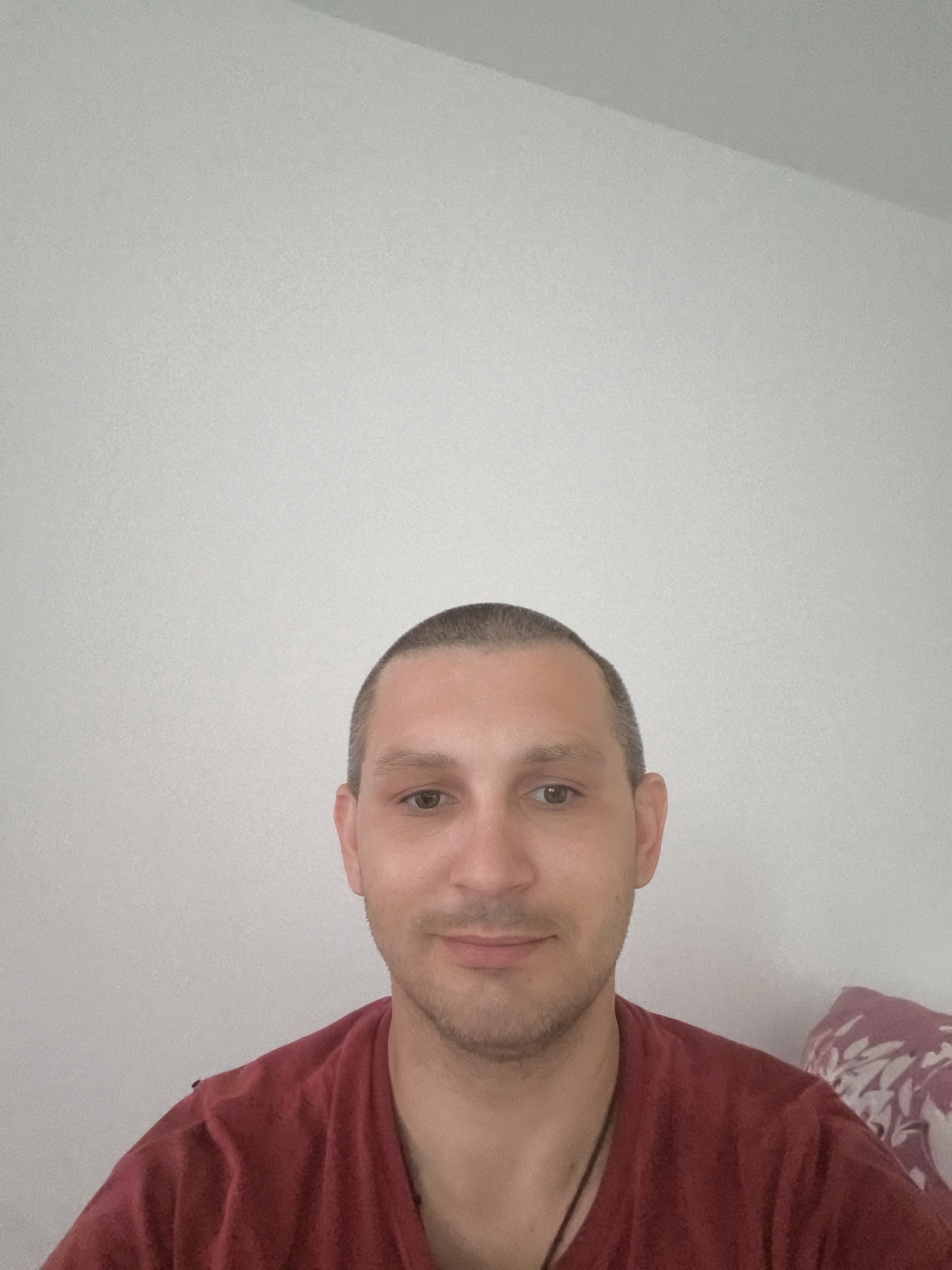 A medium shot captures a man from the chest up, with a plain white wall as the background. He has short, dark hair and is wearing a red t-shirt. The man is looking directly at the camera with a neutral expression. A small portion of a patterned pillow is visible on the right side of the image. The lighting appears to be natural, coming from the front. There are no discernible elements in the image that would indicate a specific location within Wolgast, Germany.