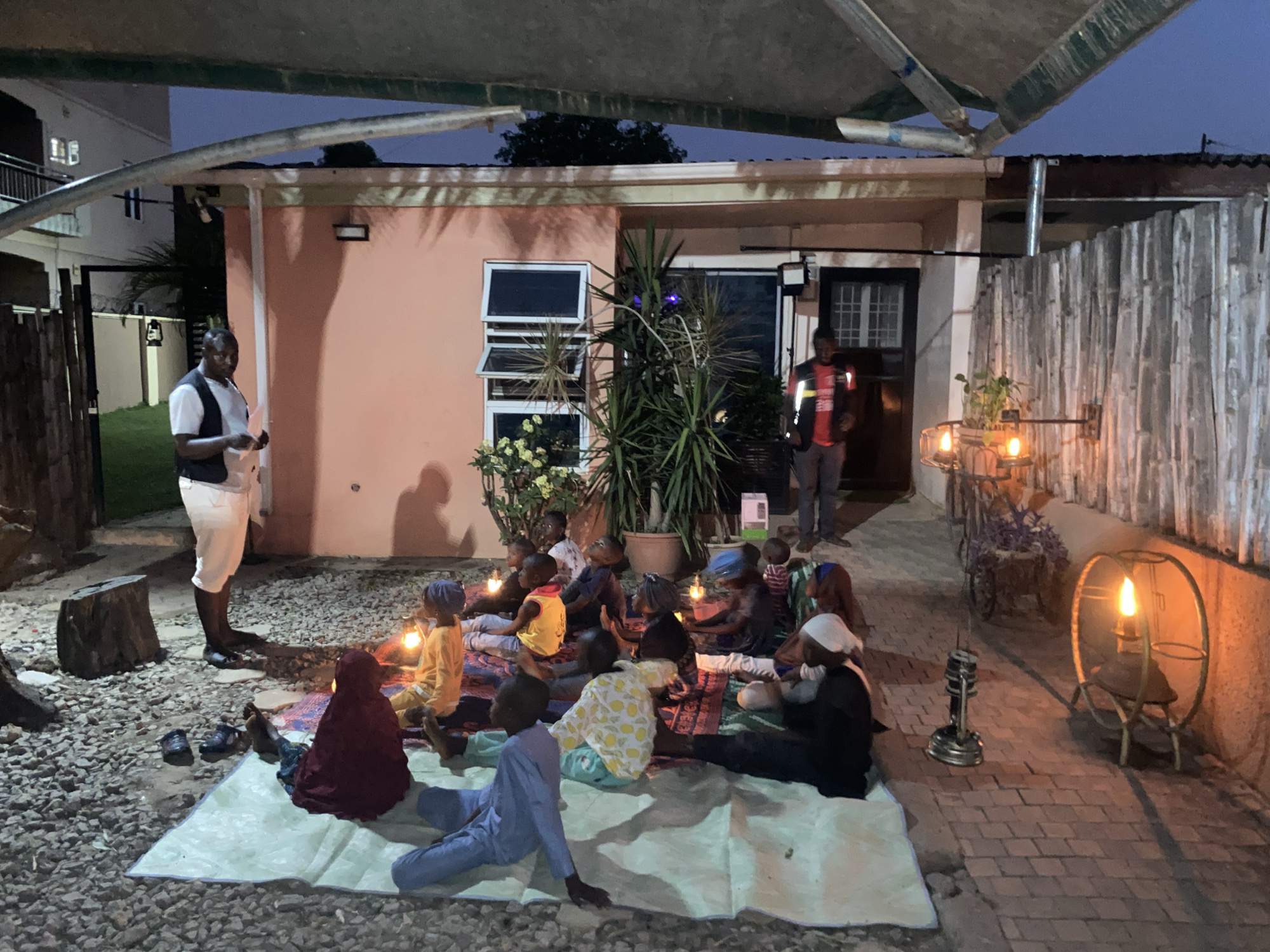In Barden Yana, Nigeria, a group of children are gathered outdoors on a light-colored mat, illuminated by individual lanterns and wall-mounted lights. They are dressed in a variety of casual clothes, some with head coverings. Two men stand nearby, one to the left wearing white pants and a dark vest, and another further back wearing a red shirt and dark vest. The setting includes a peach-colored building, plants, and a fenced perimeter. The ground is a mix of gravel and brick. An awning covers part of the gathering area.