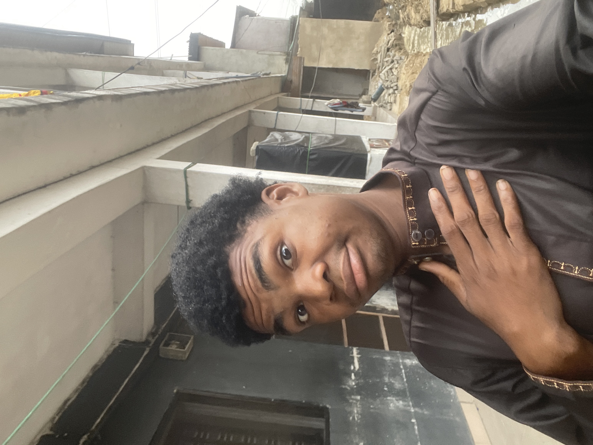 In Ado, Nigeria, a young man with dark, curly hair and brown skin takes a selfie outdoors. He is wearing a dark brown garment with gold trim around the collar and cuffs. His right hand is placed on his chest, and his left hand is not visible. His facial expression is one of mild surprise or confusion, with his eyes wide open and his lips slightly pursed. Behind him, a building with concrete columns and an unfinished upper level is visible. The ground appears to be a mix of dirt and rubble, with some water channels or trenches. The overall scene suggests an urban environment undergoing construction or in a state of disrepair.