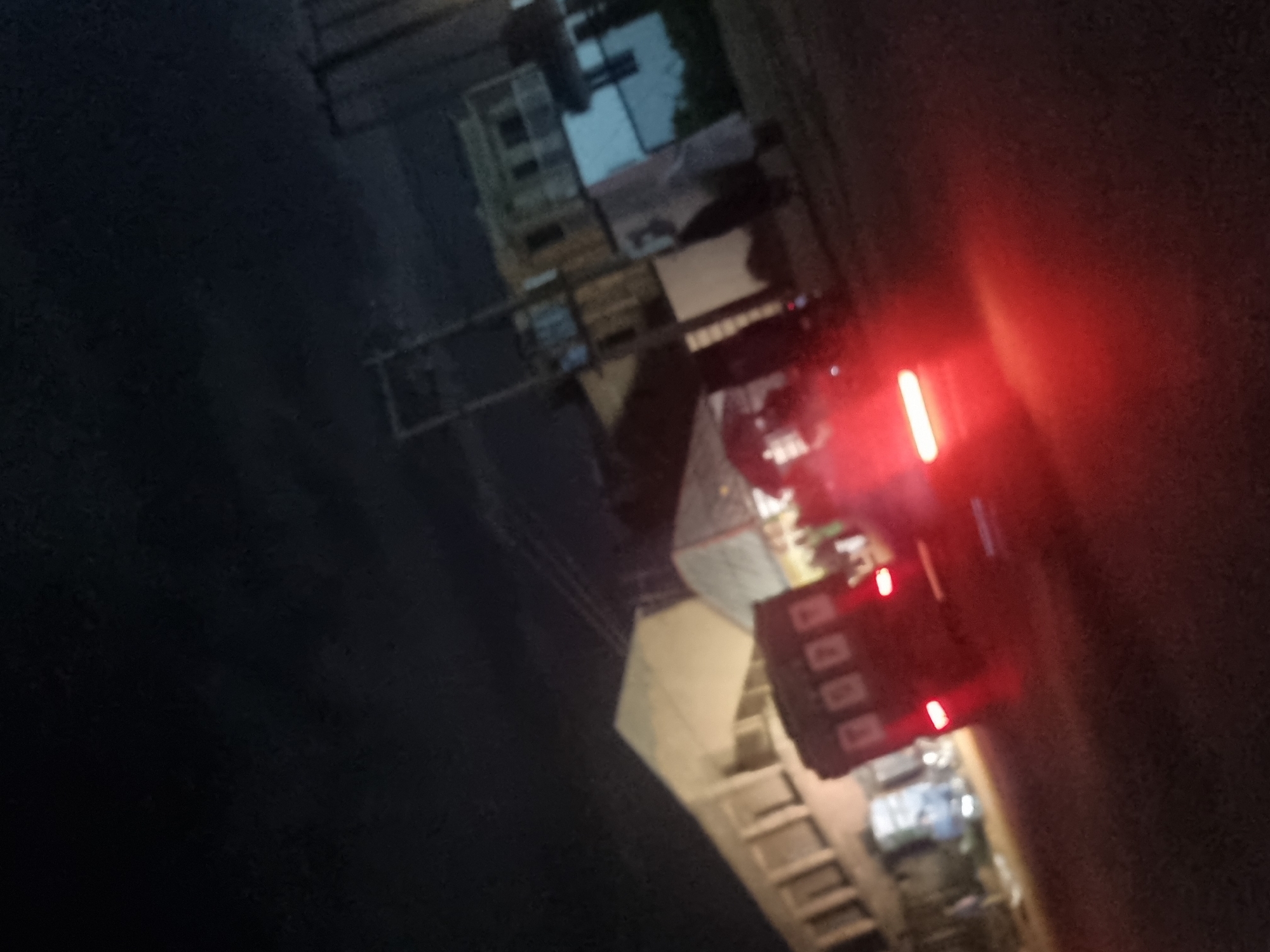 The image depicts a night scene in Gwong, Nigeria, with a focus on a red three-wheeled vehicle, possibly a tuk-tuk or a rickshaw, in motion. The vehicle's brake lights are illuminated, casting a strong red glow on the dark, possibly unpaved, road surface. Behind the vehicle, a long, light-colored building with multiple windows is visible, suggesting a commercial or residential area. Further in the background, several other structures, including a multi-story building with white and light-colored facades and darker rooflines, are discernible. Power lines and poles are also present in the background, indicating the presence of infrastructure. The sky is dark, suggesting nighttime or overcast conditions. The overall impression is of a busy street scene at night, with the illuminated vehicle being the primary subject.