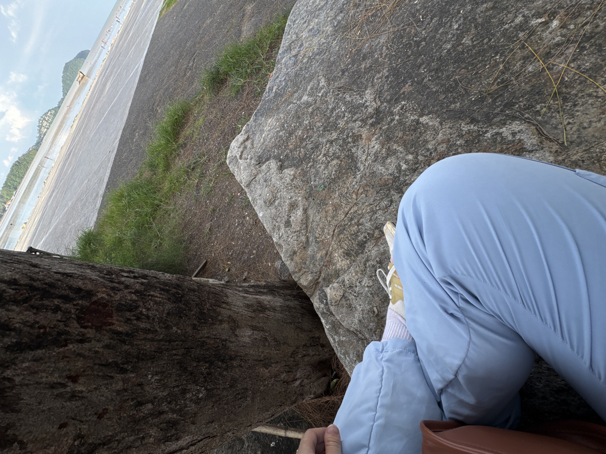 A person is sitting on a rock next to a tree trunk, wearing light blue pants and white and yellow sneakers with pink socks. They are in an outdoor setting with a paved area that slopes down towards a body of water. Hills and some buildings are visible in the distance. The sky is partly cloudy. The scene appears to be a public area near a waterfront, possibly a park or a scenic overlook in Phuket City Municipality, Thailand.