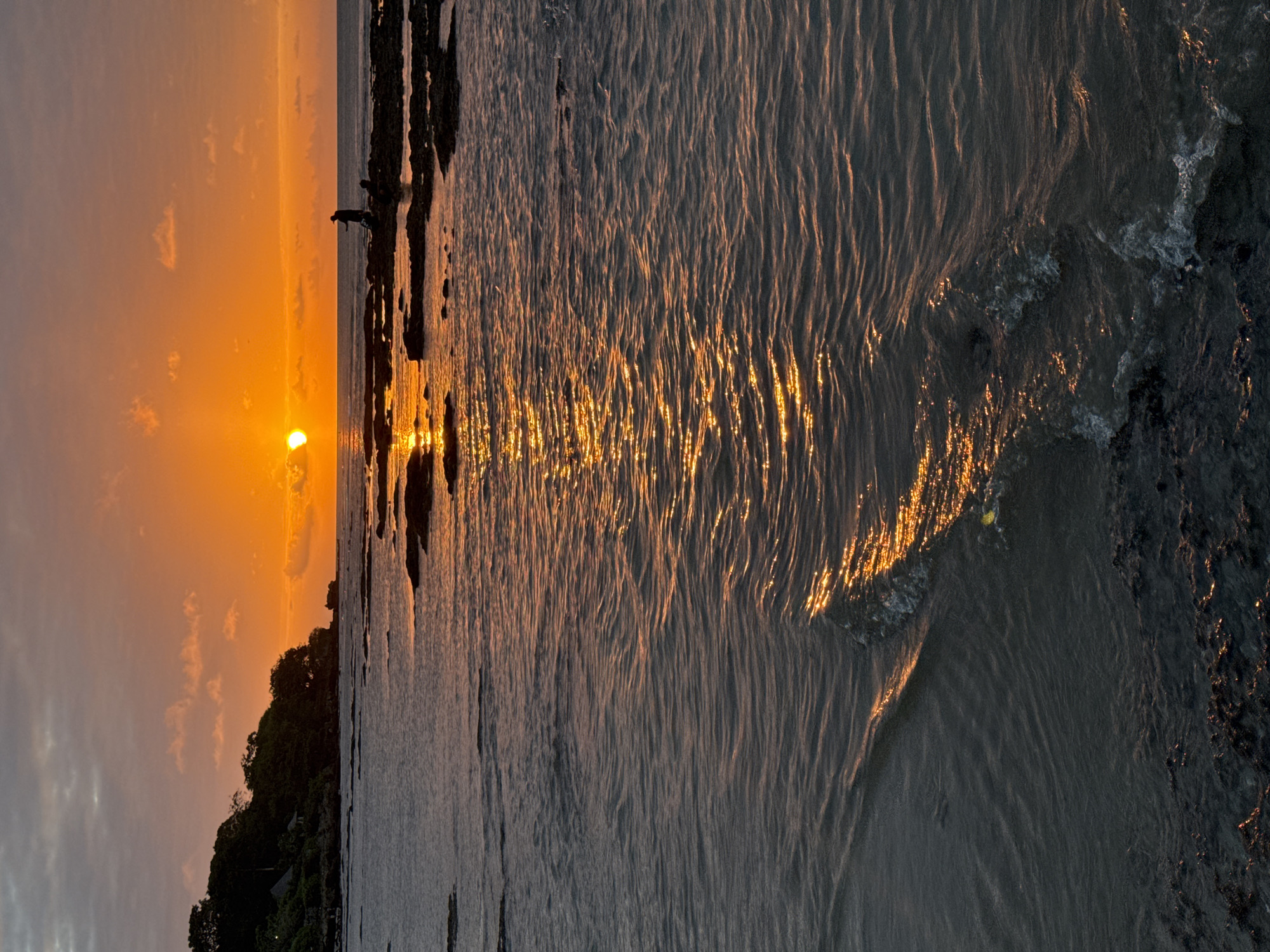An outdoor, vertical, eye-level shot shows a tranquil ocean scene at sunset. The sun is a bright orange orb on the horizon, casting a golden reflection across the rippling water. The ocean is calm, with small waves lapping at a rocky shore. A lone figure stands on the rocks in the distance, silhouetted against the bright sky. The shore is lined with trees and a few buildings. The image was taken in Kuta Selatan, Bali, Indonesia, a popular tourist destination known for its beautiful beaches and sunsets.